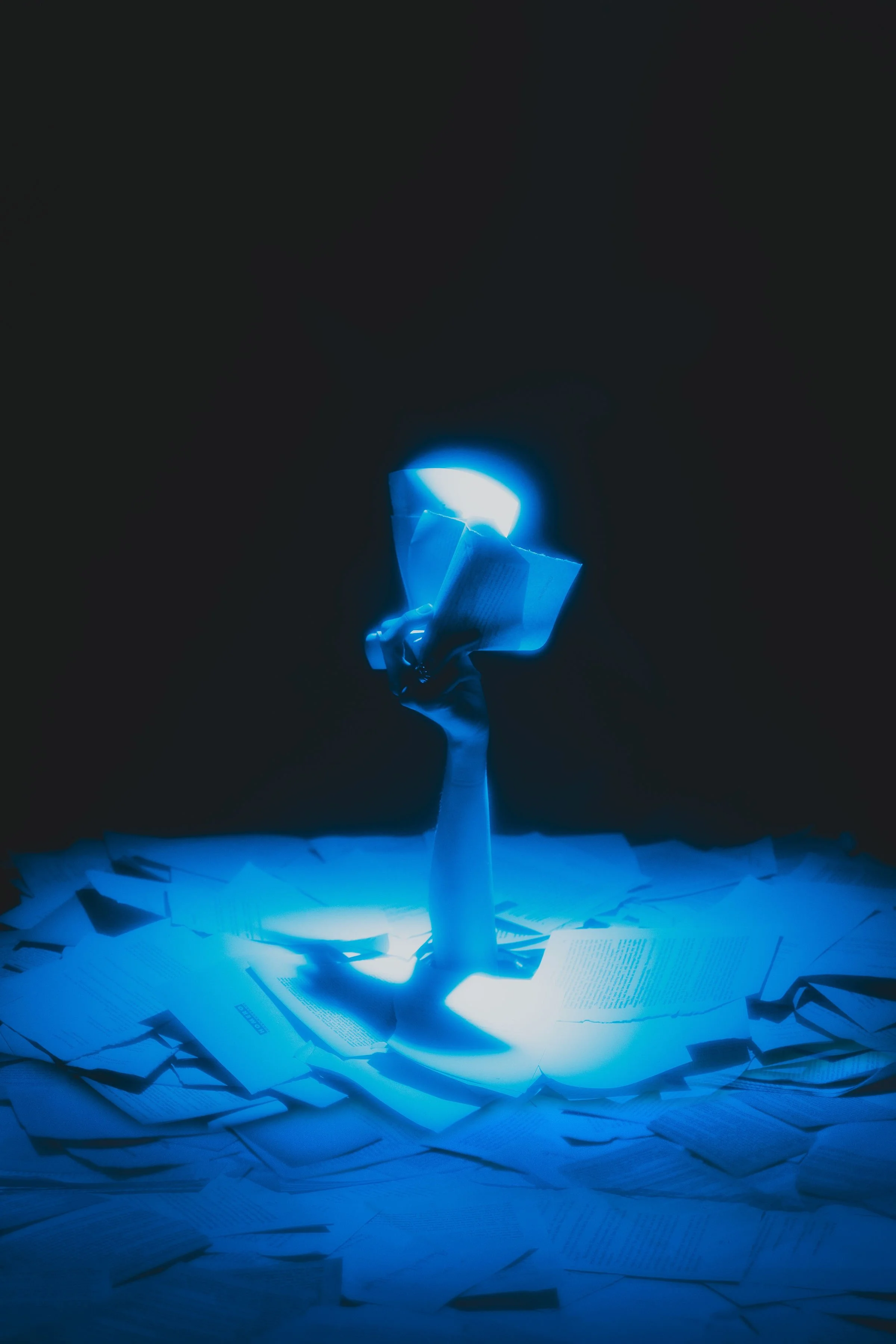 caitlin kelly Hand emerging from a pile of scattered papers, holding a lit lighter with a bright flame illuminating the dark background.