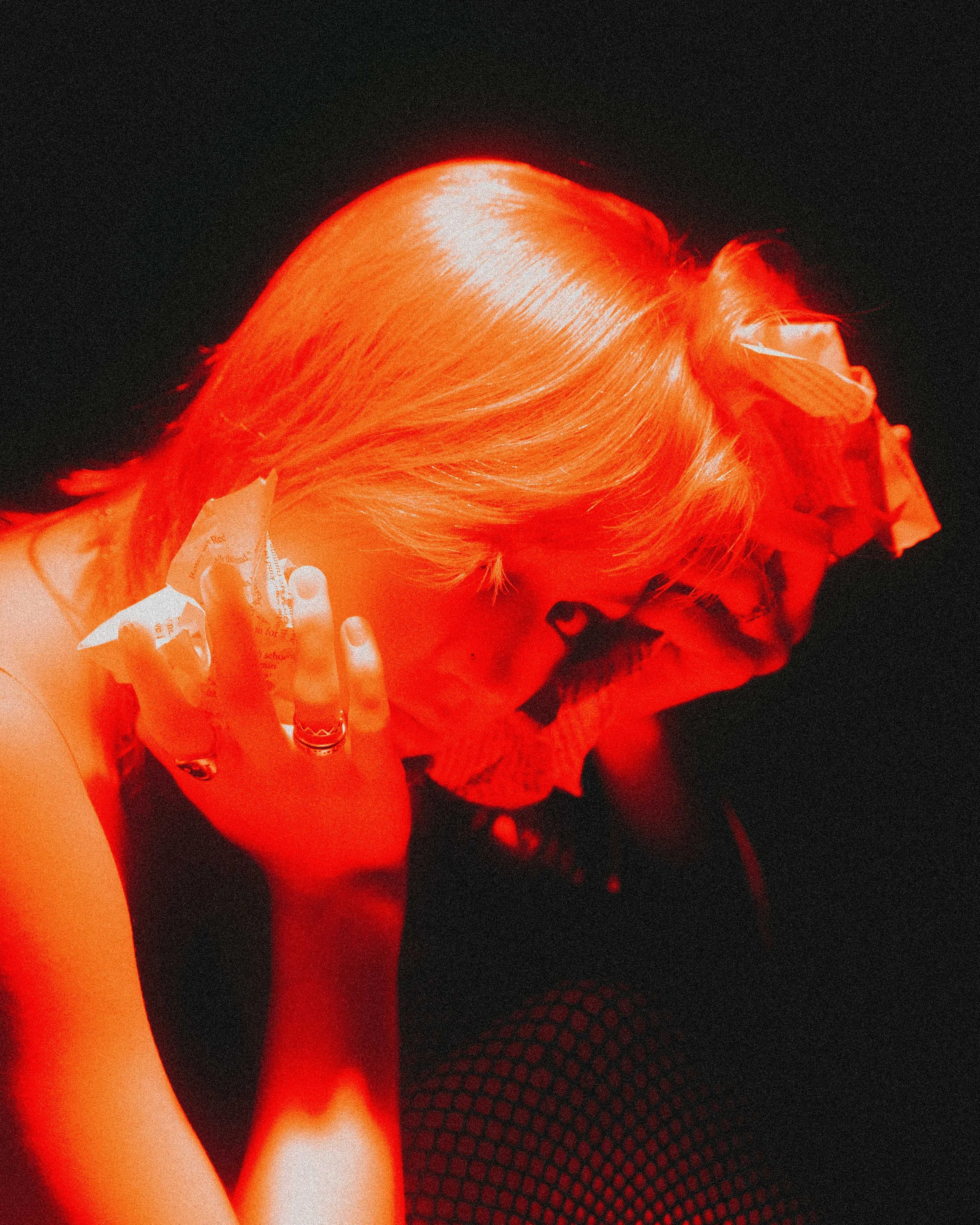 A woman with blonde hair holding a torn piece of paper to her face, with fishnet tights visible and a black background.