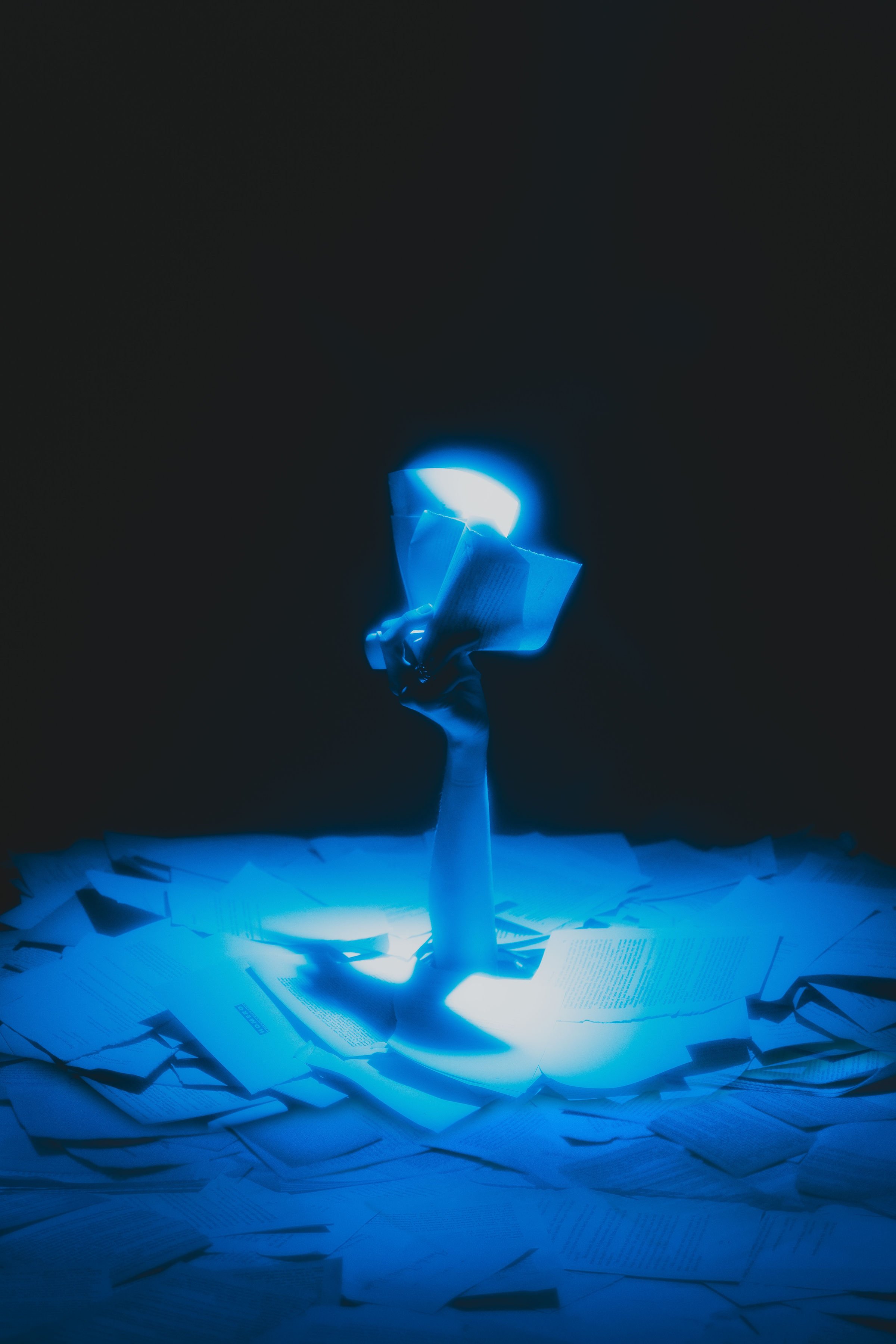 A hand holding a computer mouse emerging from a pile of scattered papers, illuminated by a blue light in a dark environment.