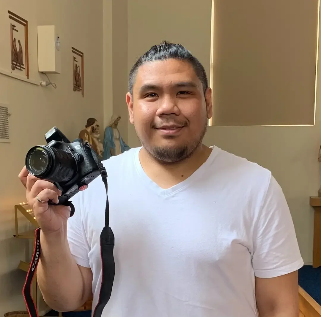 Man smiling and holding a compact DSLR camera with a strap in a room with religious statues and framed artwork.