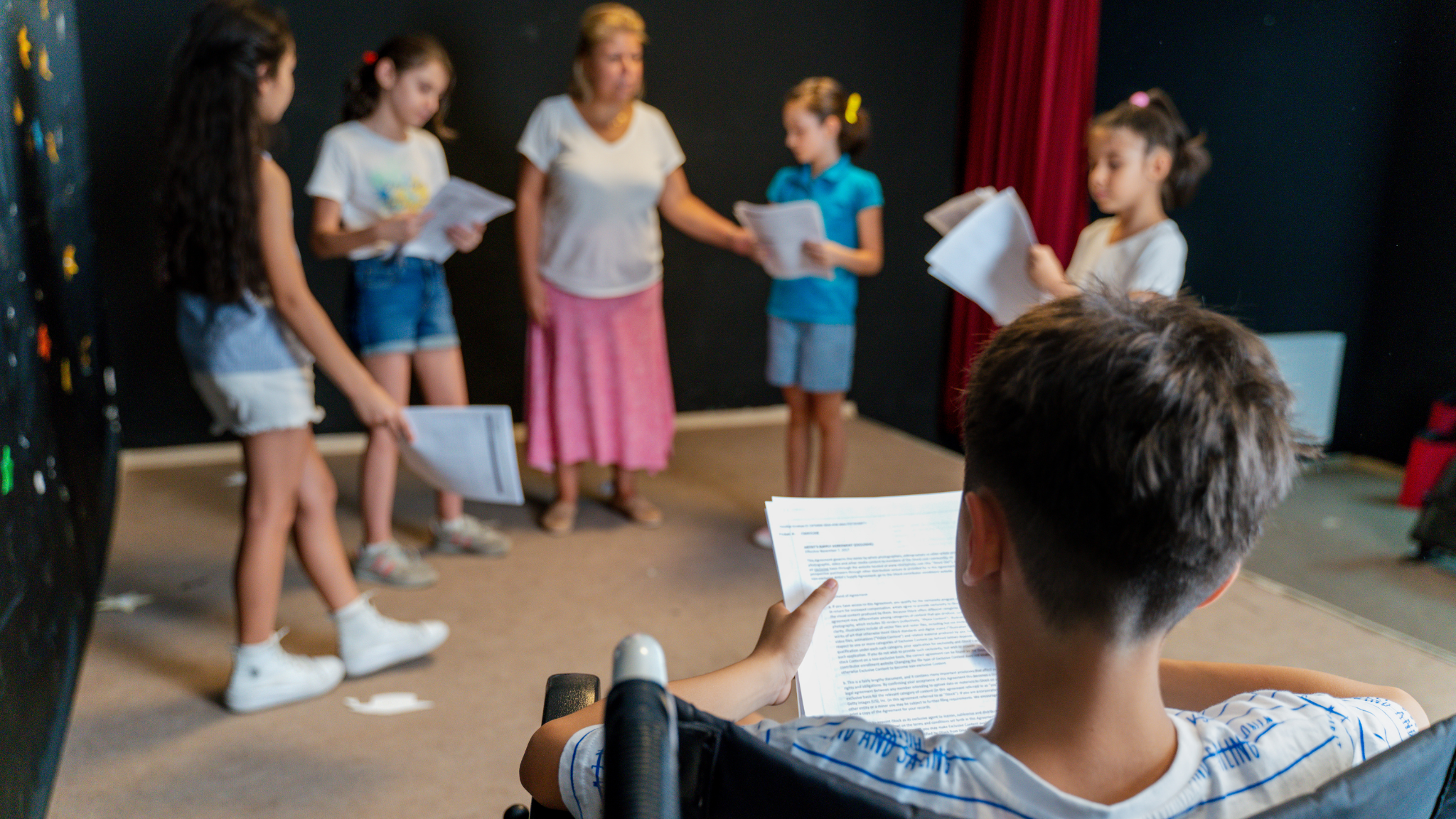 Children and a woman standing in a room, holding scripts or papers, likely rehearsing or practicing a play or presentation.