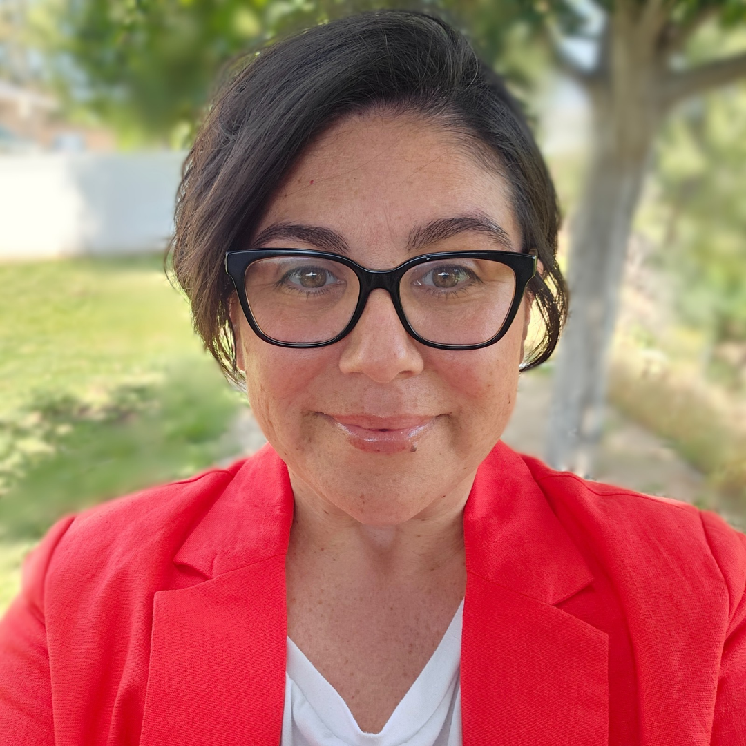 A woman wearing glasses, a red blazer, and a white shirt, smiling outdoors with trees and greenery in the background.