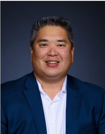 Portrait of a man with short, dark hair wearing a dark blue blazer and white shirt, smiling against a dark background.