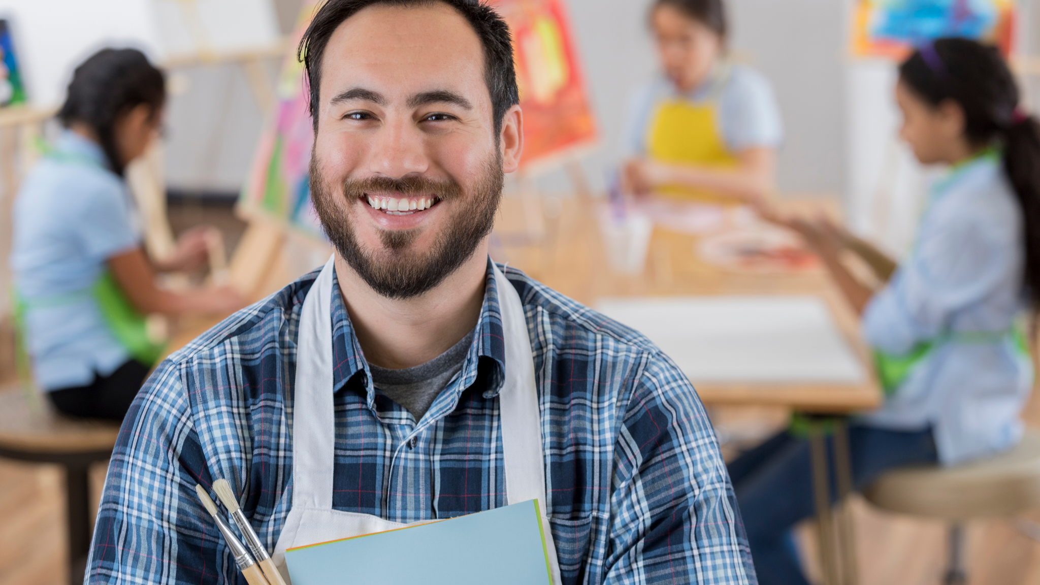 A smiling man with a beard wearing a plaid shirt and an apron, holding art supplies, in an art classroom with children painting in the background.