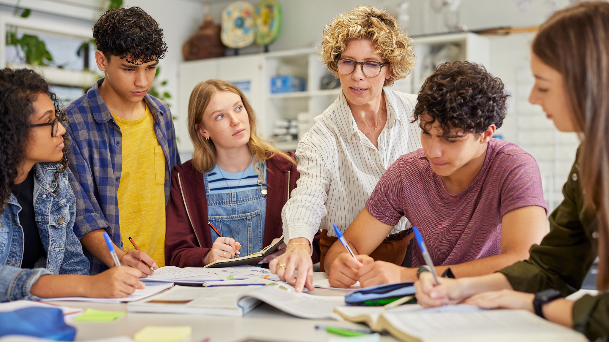 Teacher instructs a group of diverse students during a classroom lesson with open books and notebooks on table.