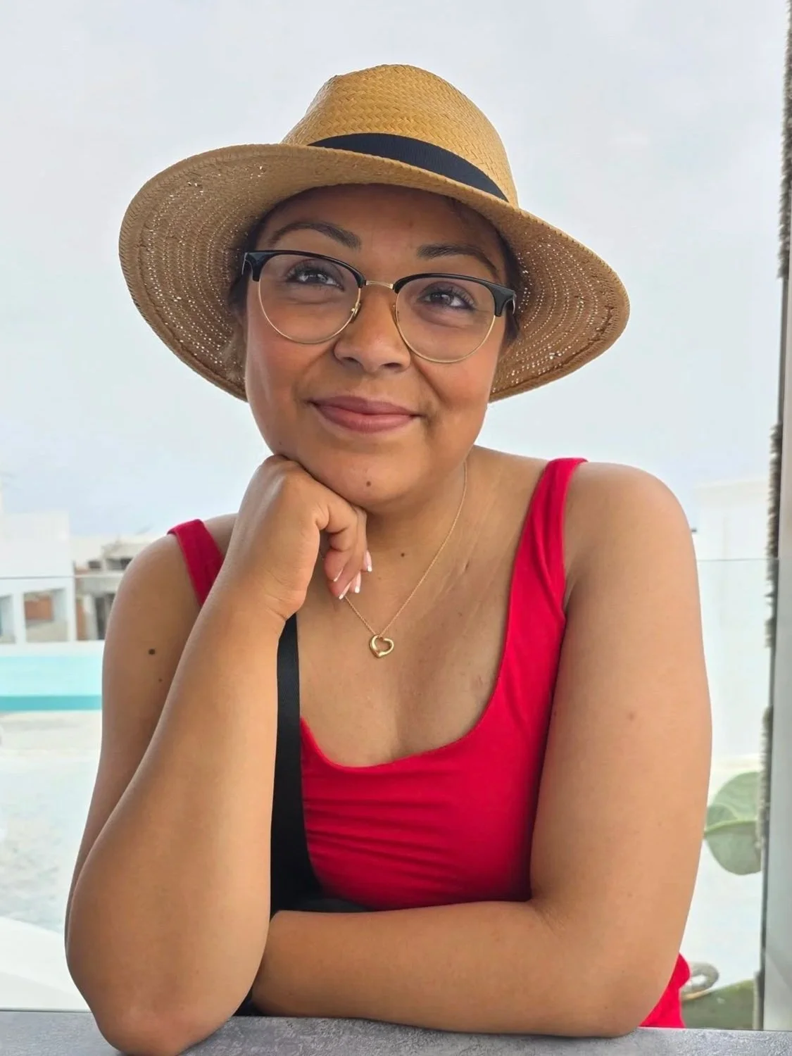 A woman wearing glasses and a large straw sunhat, resting her chin on her hand, with a red sleeveless top and a gold necklace shaped like a heart, sitting indoors with a large window behind her.