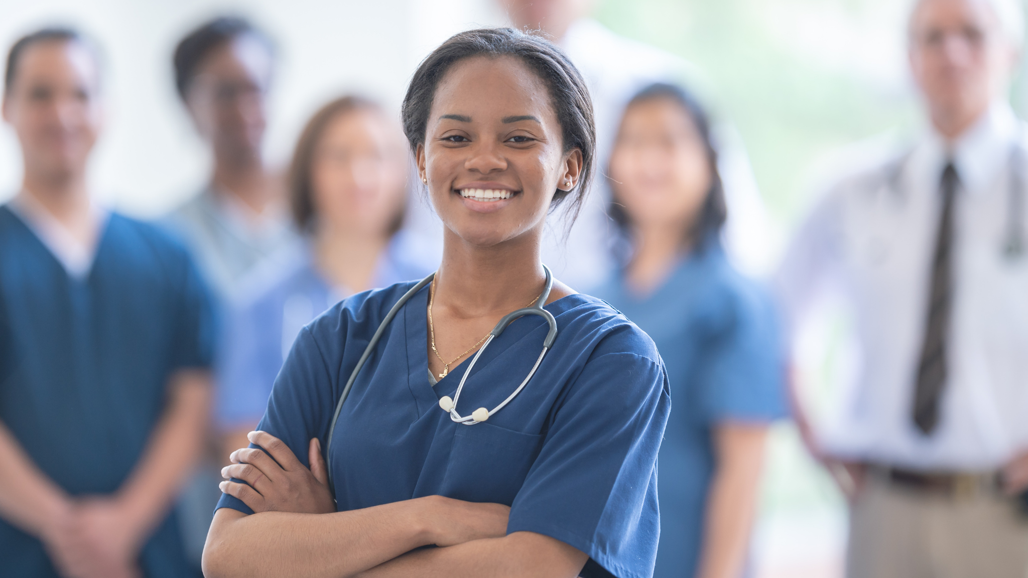 A group of healthcare professionals, including a smiling female nurse in the foreground wearing scrubs and a stethoscope, with others in medical attire blurred in the background.