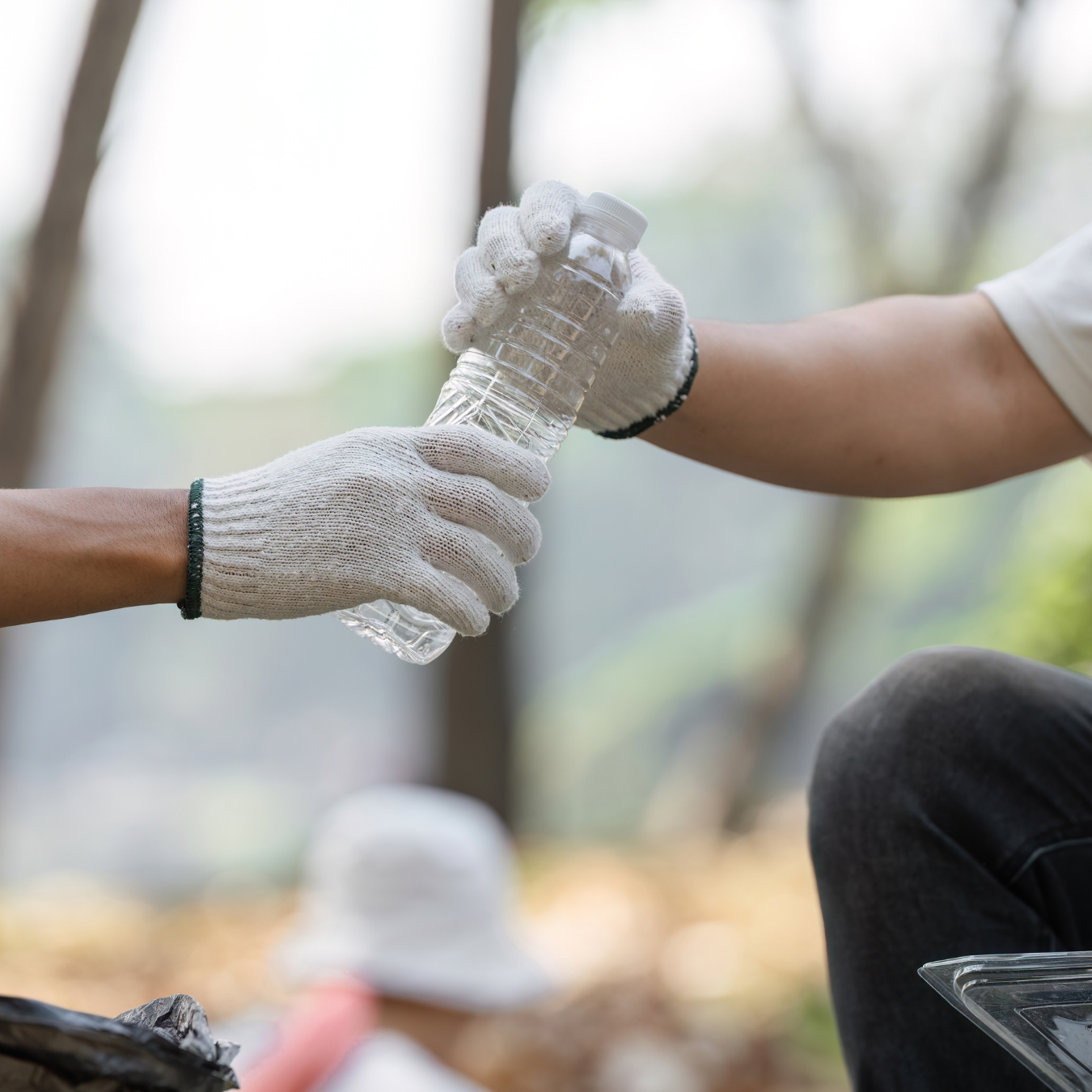 Two people exchanging a clear plastic water bottle outdoors, both wearing white gloves, with blurred background of trees and a person wearing a white hat.