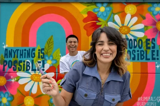 Two people smiling in front of a colorful mural with rainbow, flowers, and the text 'Anything is Possible!'