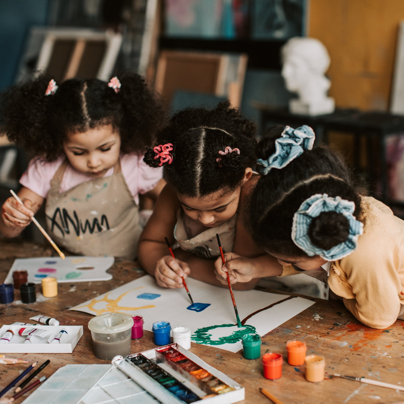 Three young girls with curly hair painting on a large sheet of paper at a table with various paint supplies, including bottles, brushes, and a palette.