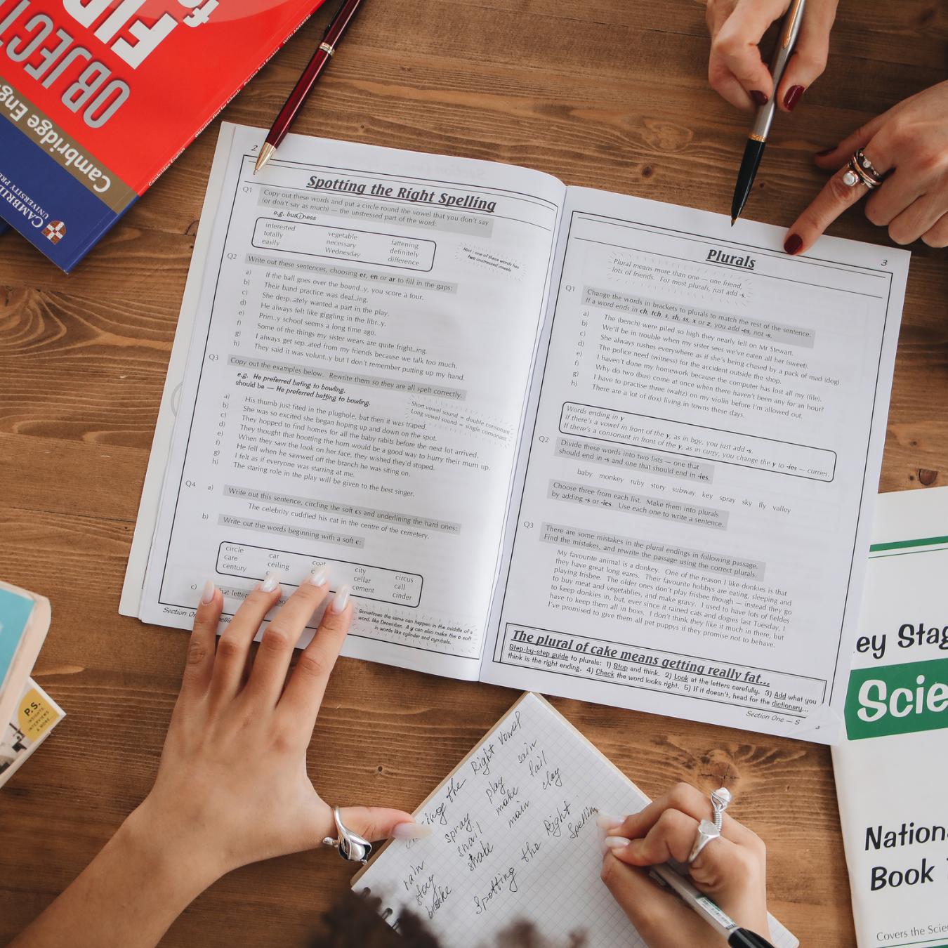 A person taking notes in a small notebook at a desk with open textbooks on English grammar and spelling, and a science textbook nearby.