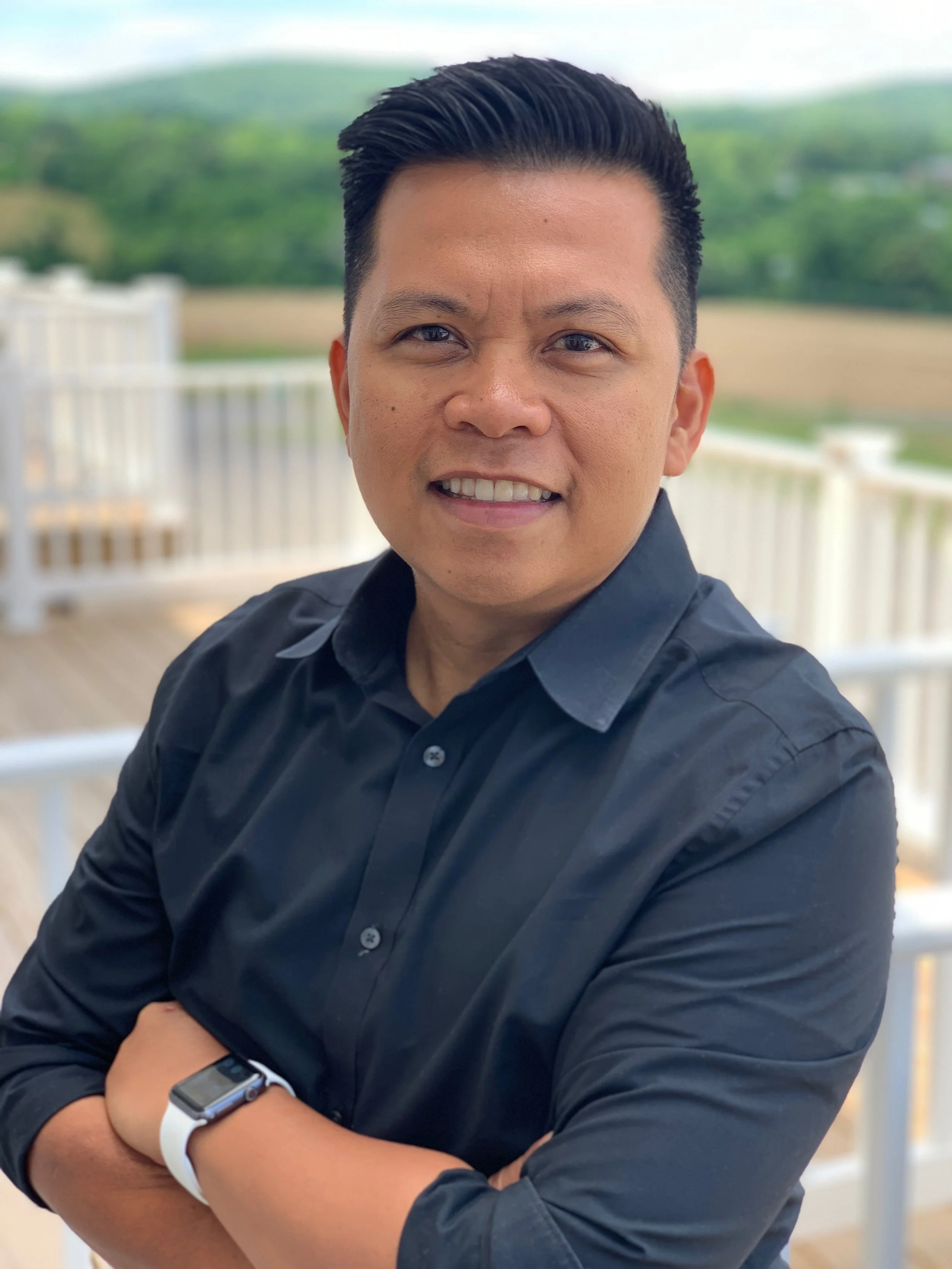 A man wearing a black shirt and a smartwatch, smiling outdoors on a balcony with a green landscape in the background.