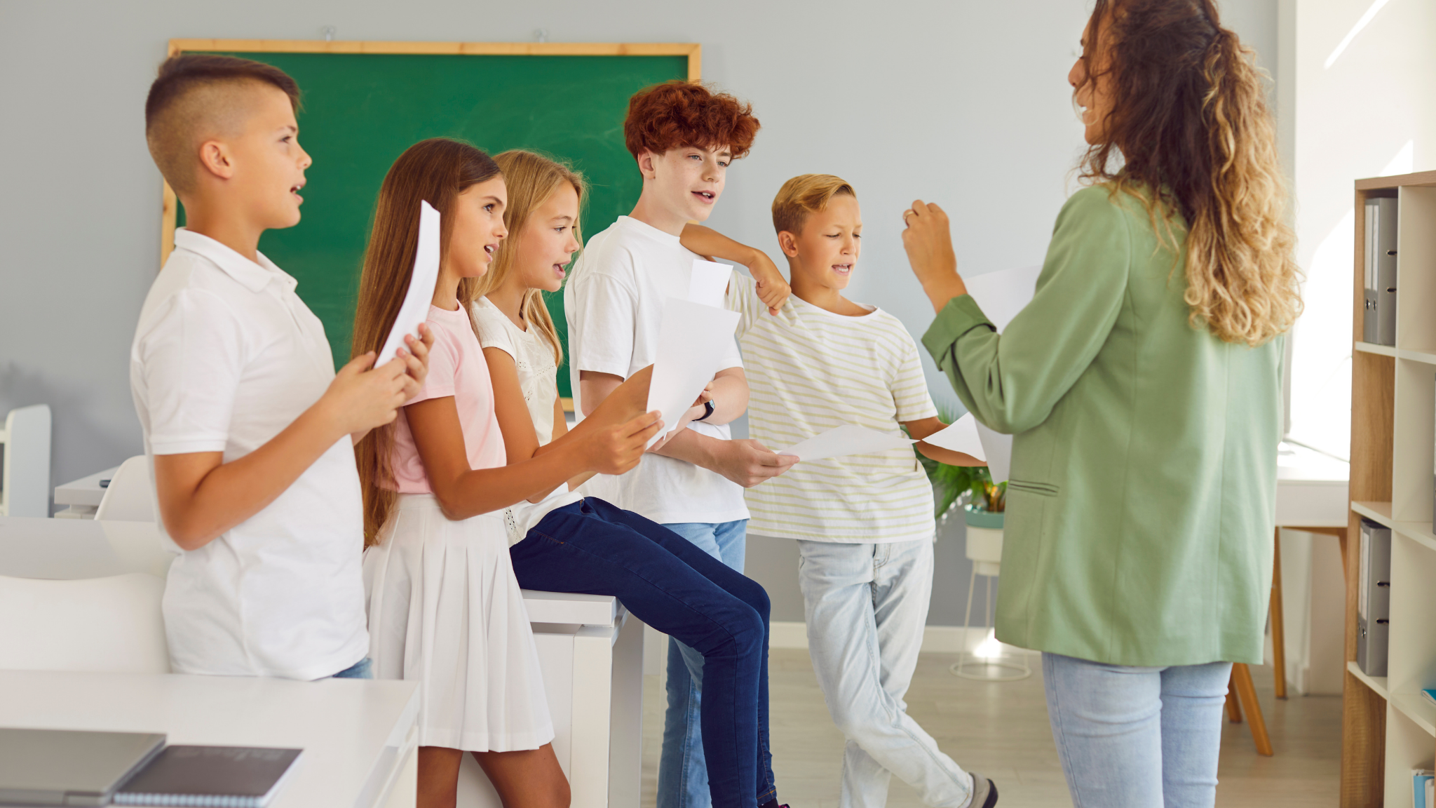 A teacher is giving instructions to five children, holding papers, in a classroom with a green chalkboard in the background.