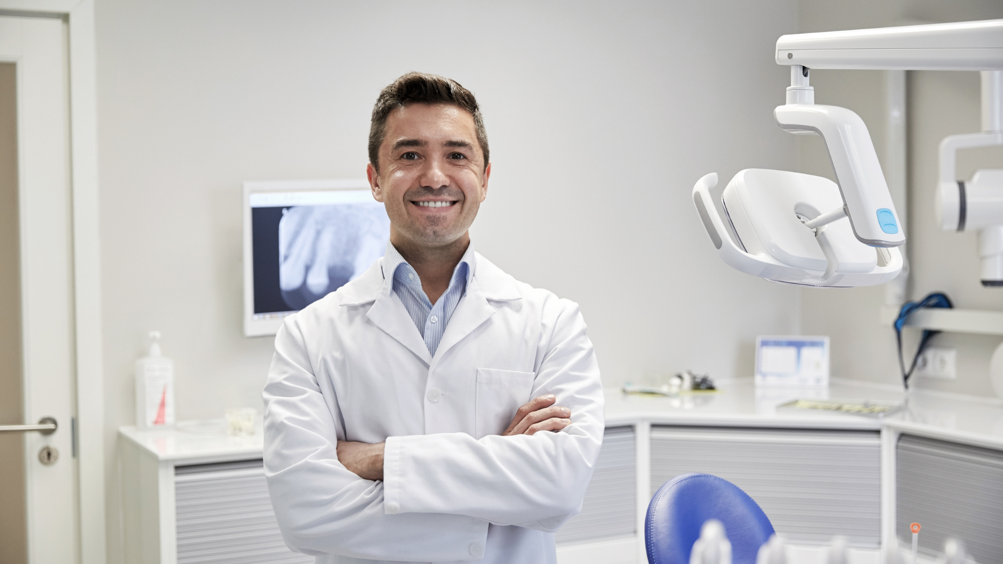 A male dentist smiling with arms crossed in a dental clinic, with dental equipment and X-ray images in the background.