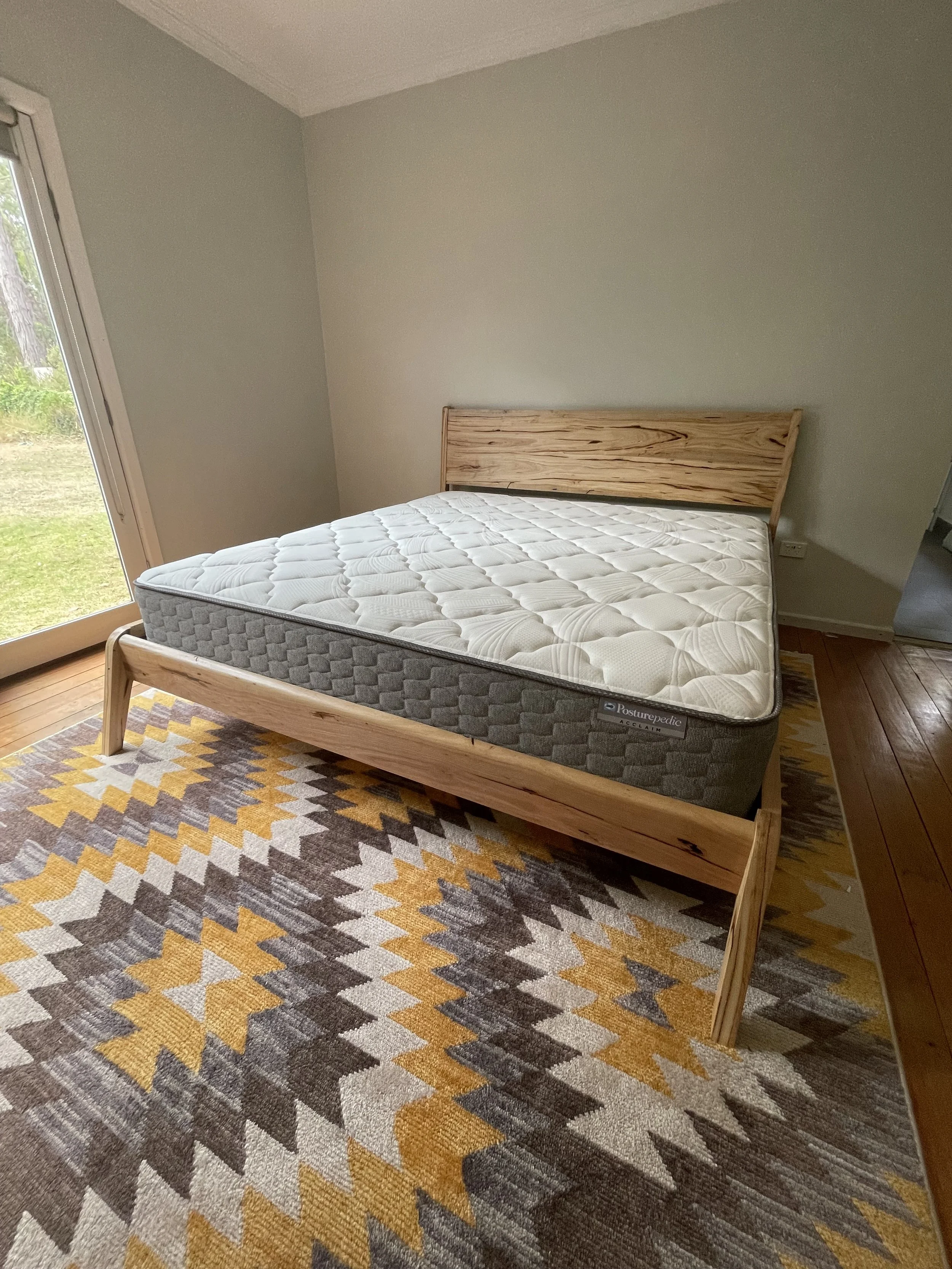 A bedroom with a wooden bed frame and a mattress, situated on a patterned area rug. A sliding door to the outside is visible on the left, allowing natural light into the room.