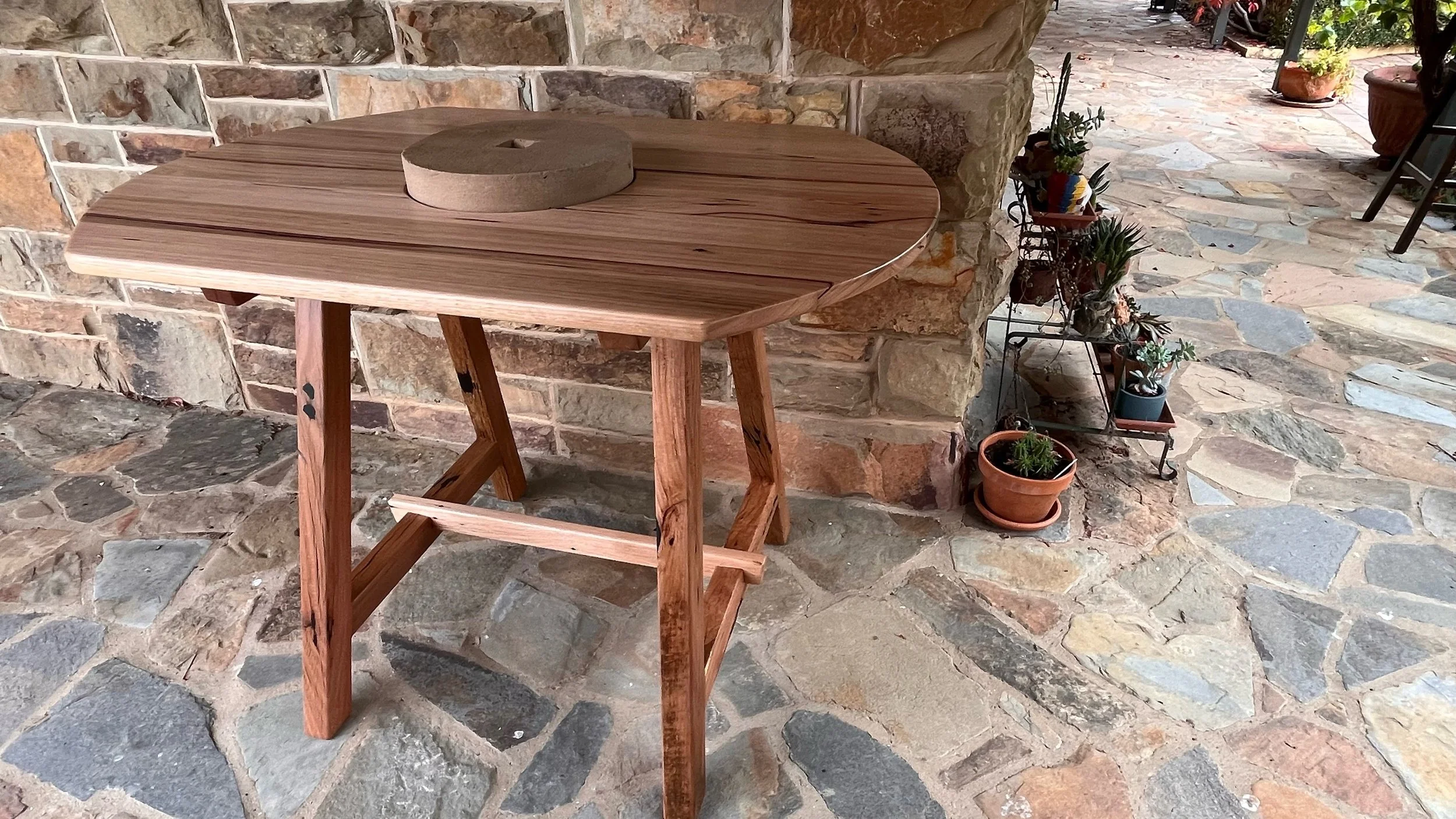 A wooden table with a circular stone object on top, set on a stone patio near a brick wall and potted plants.