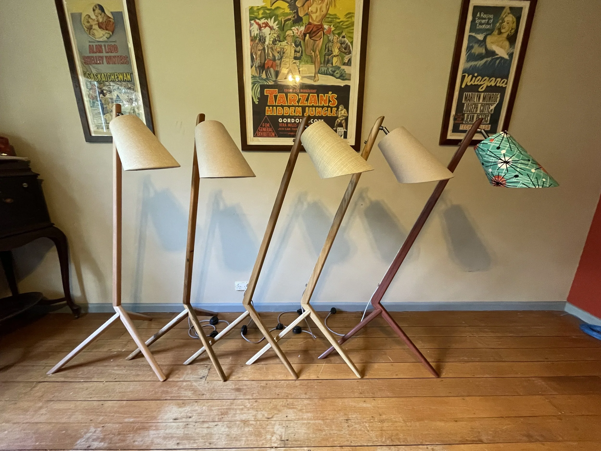 Five vintage style floor lamps with tripod wooden stands and fabric shade heads, one with a green patterned shade, lined up against a wall in a room.