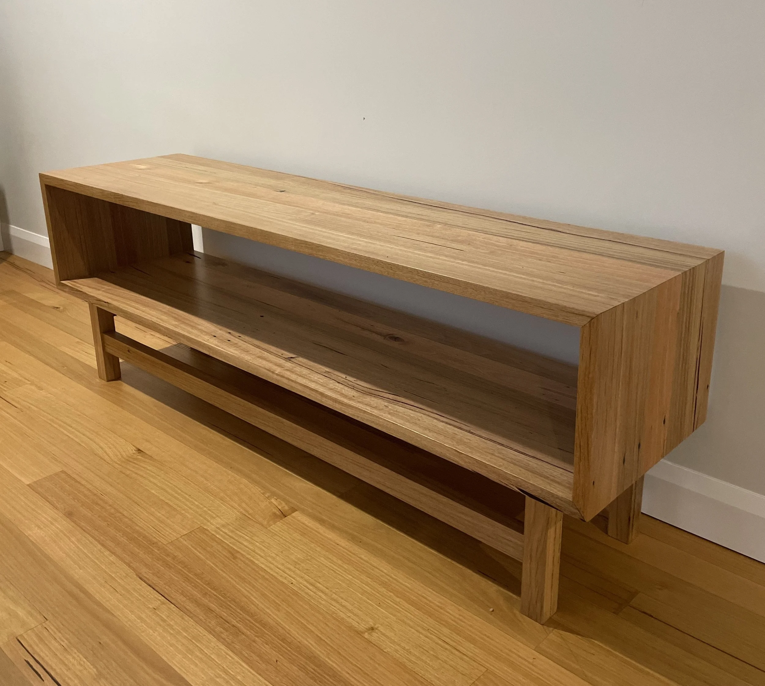 A wooden console table with open shelves placed against a white wall on a hardwood floor.