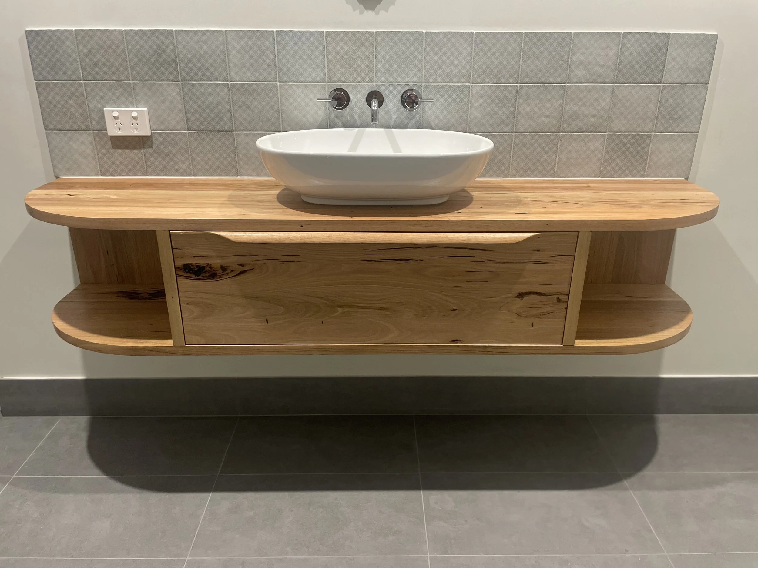 Wooden floating vanity with a white oval vessel sink and dual faucet fixtures, tiled backsplash, electrical outlet, and gray floor tiles.