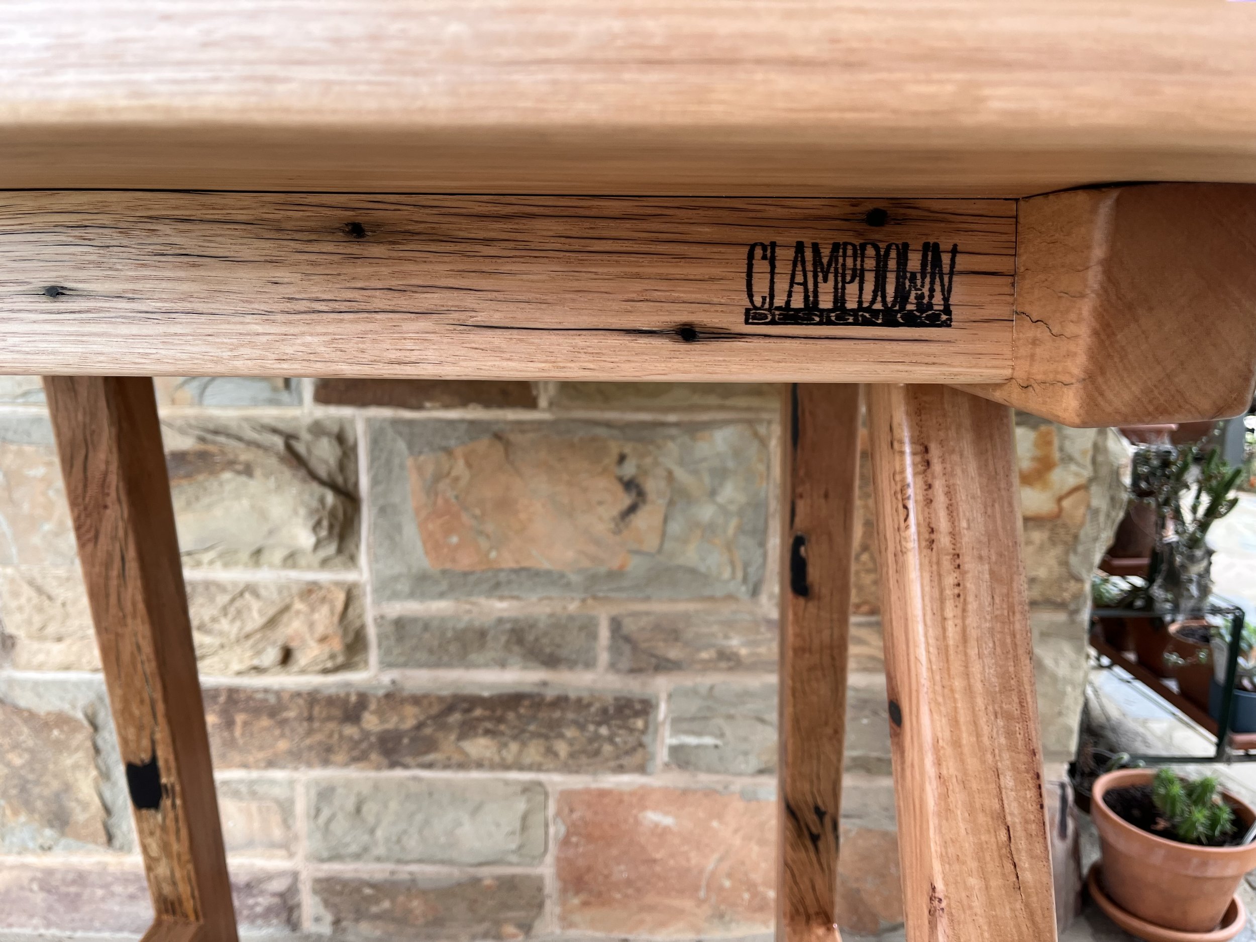 Close-up of a wooden furniture piece with 'CLAMPDOWN DESIGNS' logo and a brick wall background.