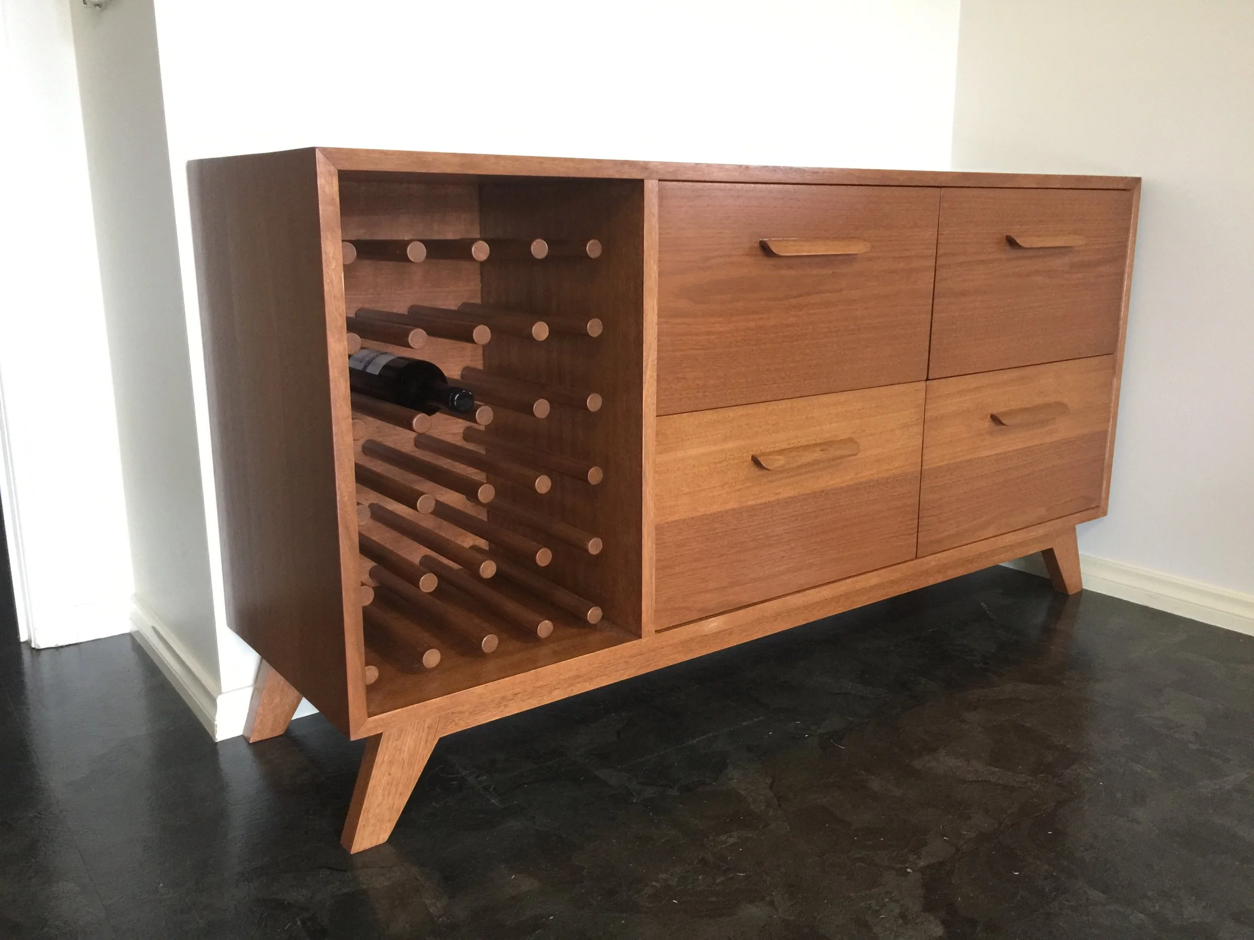 A wooden sideboard with four drawers and angled legs, featuring an open wine rack on the left side that holds a single wine bottle.