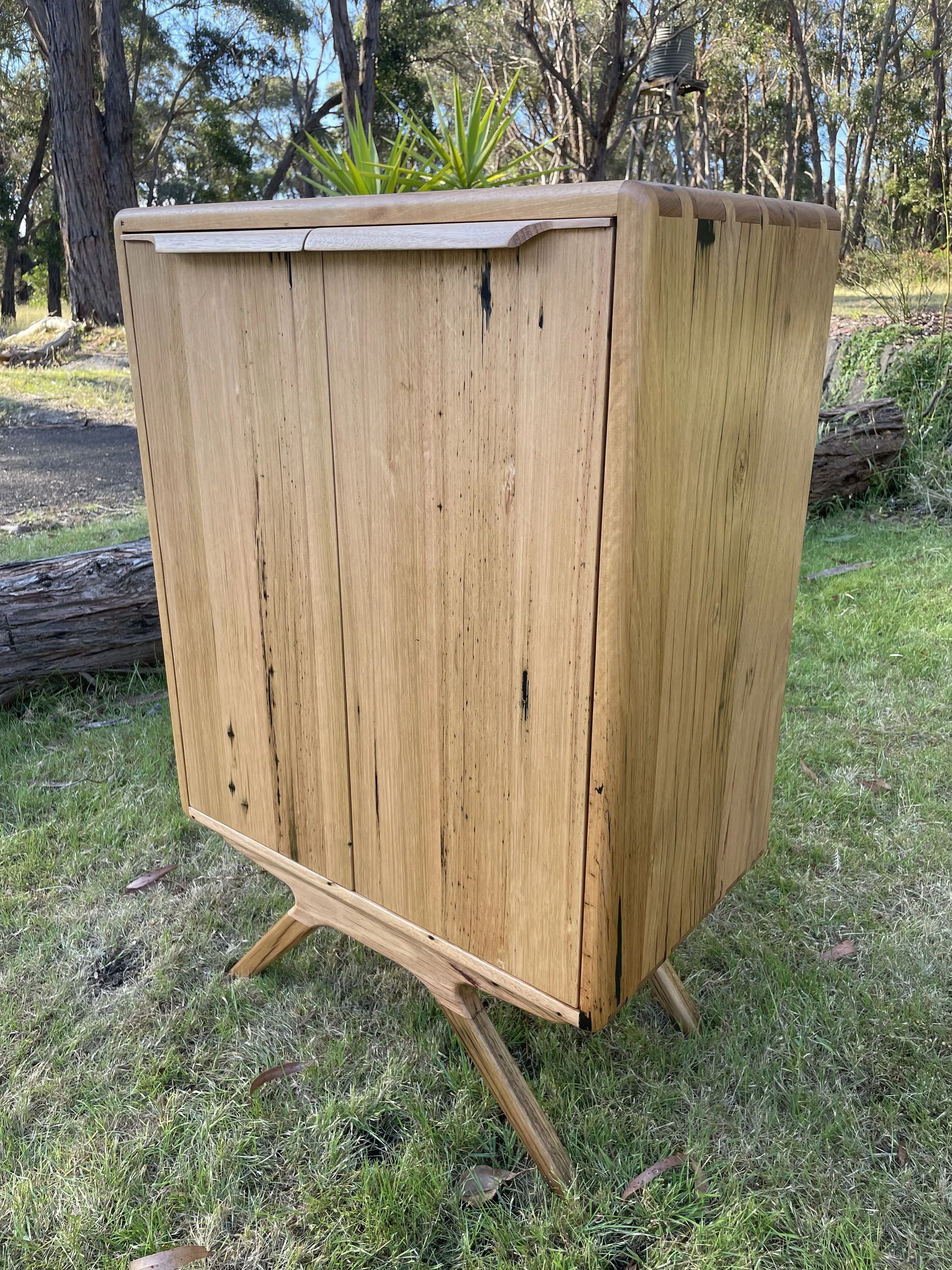 A wooden cabinet with a plant on top, situated outdoors on grass, with trees and a birdhouse in the background.