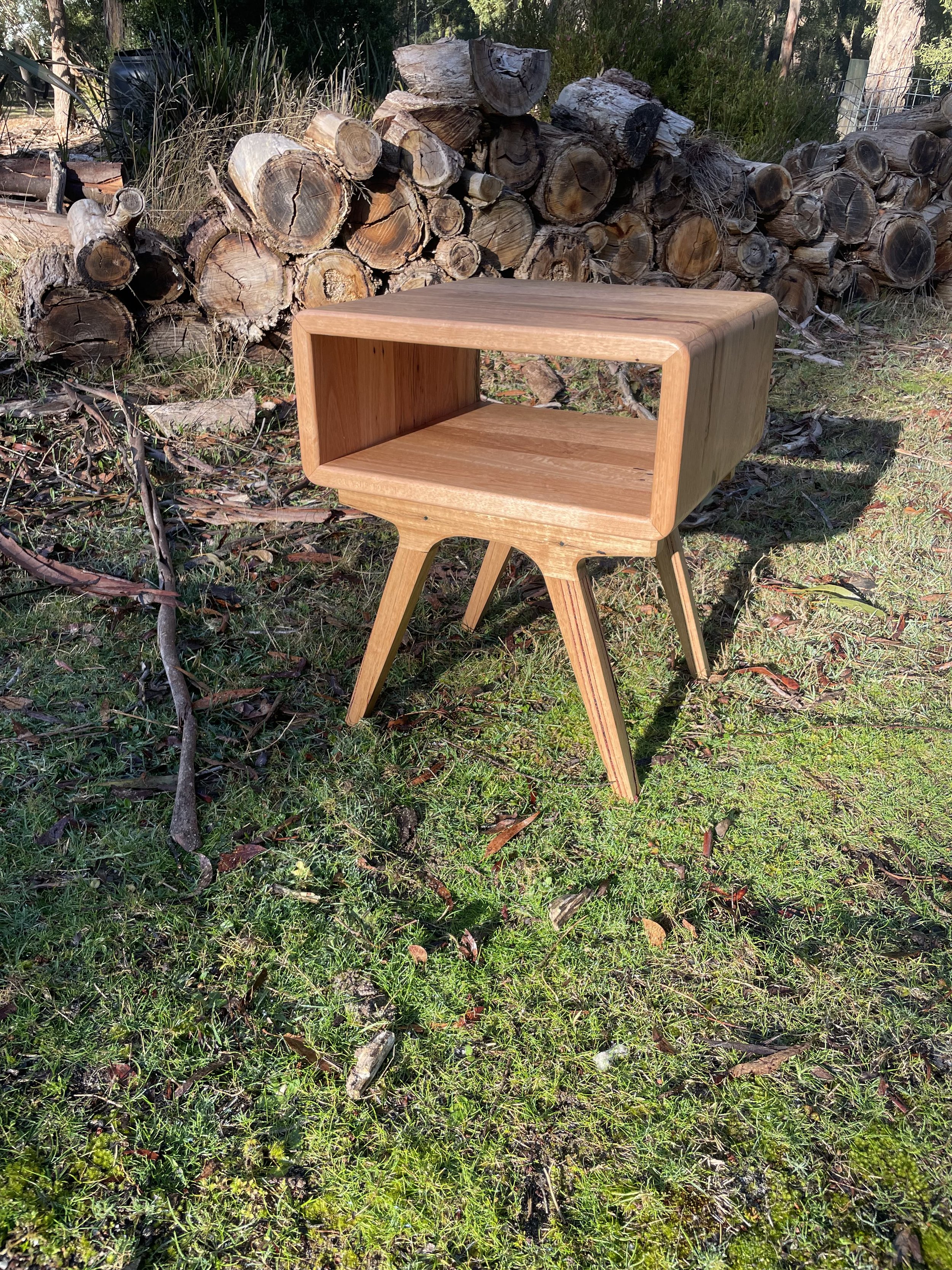 A wooden side table outdoors on grass, with a background of stacked logs and trees.
