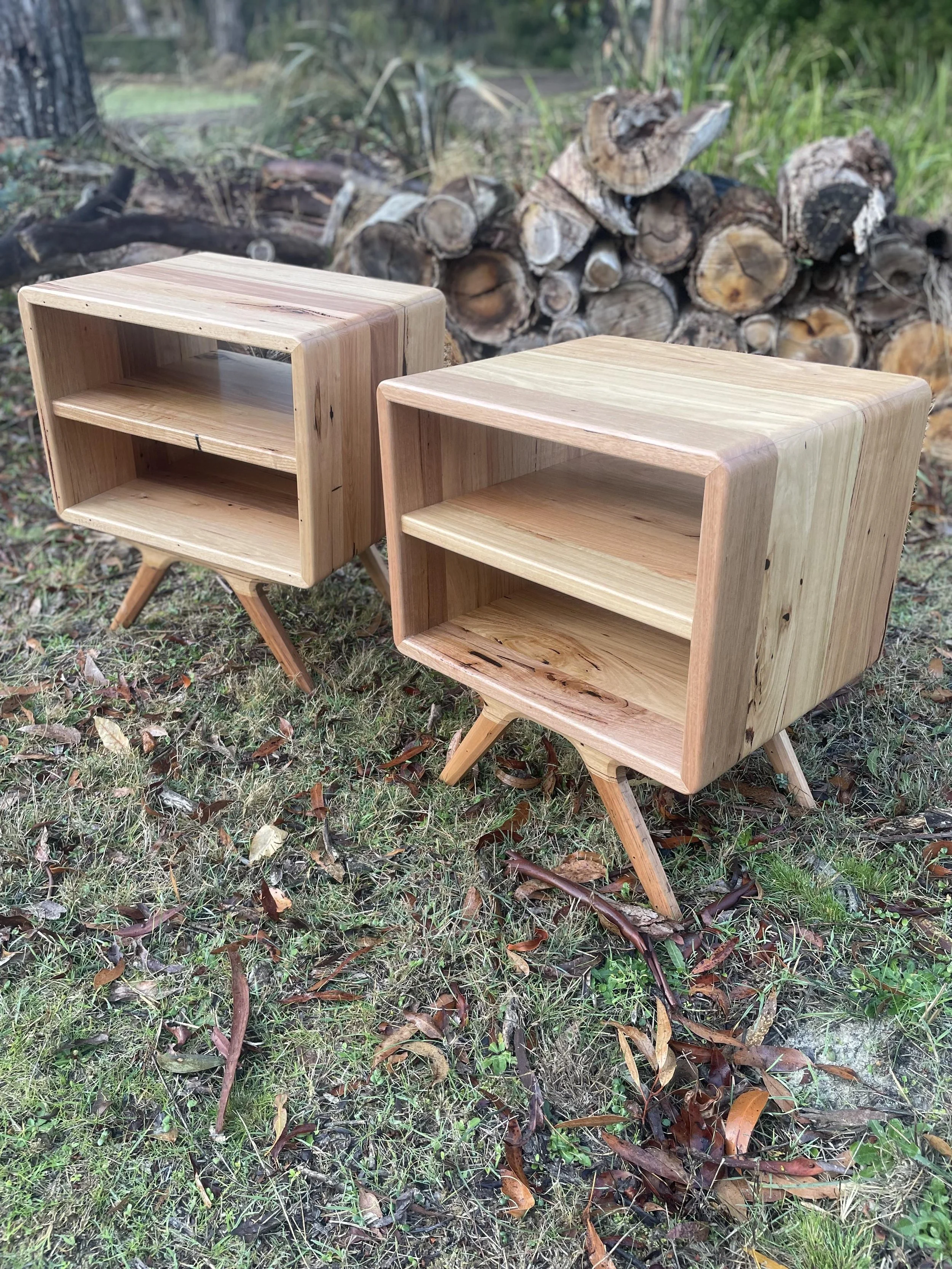 Two wooden nightstands with angled legs positioned outdoors on grass with fallen leaves, set in front of a pile of cut firewood in a forested area.