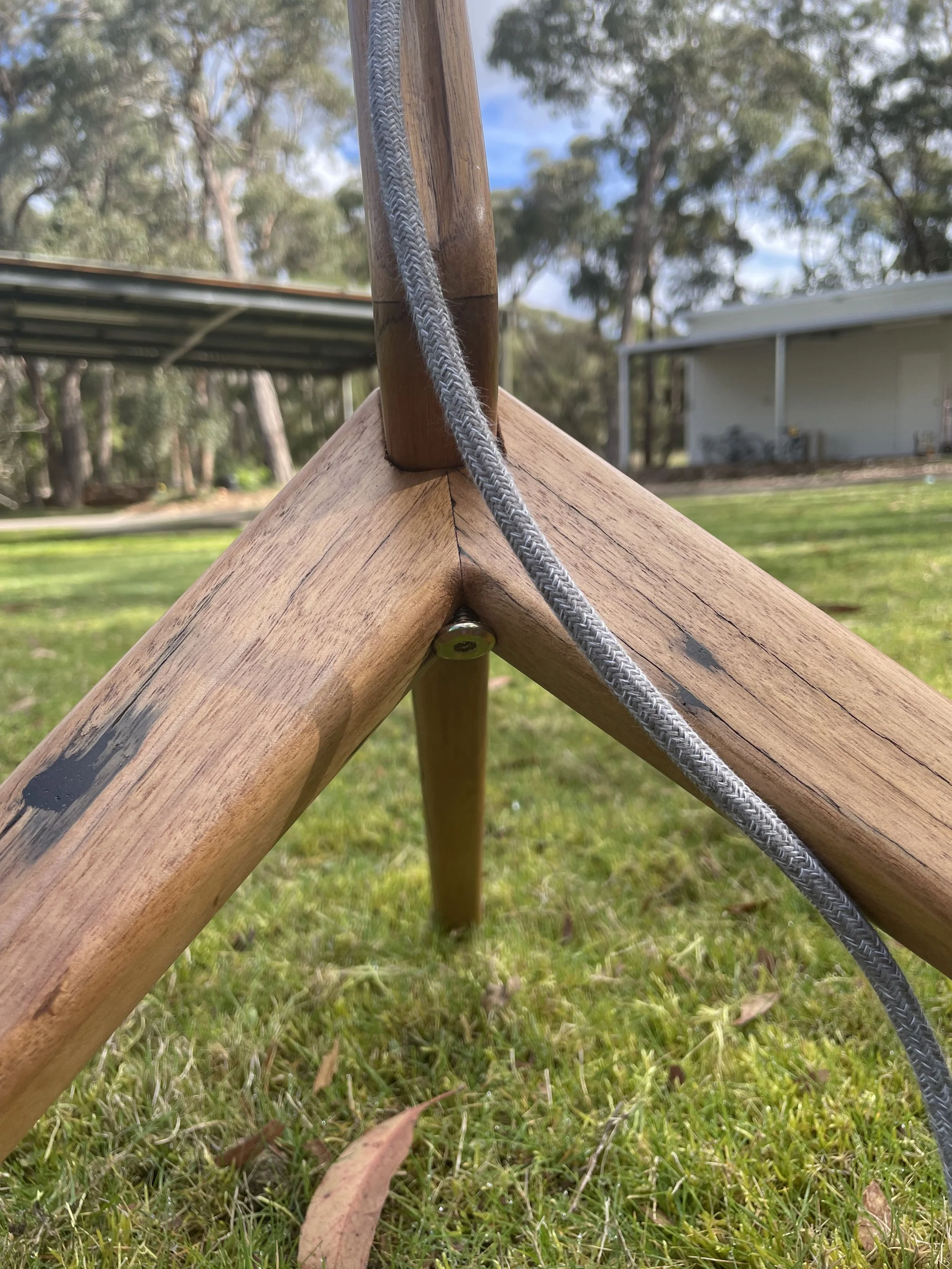 Close-up of a wooden structure, possibly a piece of outdoor furniture or playground equipment, with a metal bolt and a gray braided rope hanging over it, on a grassy lawn with trees and a building in the background.