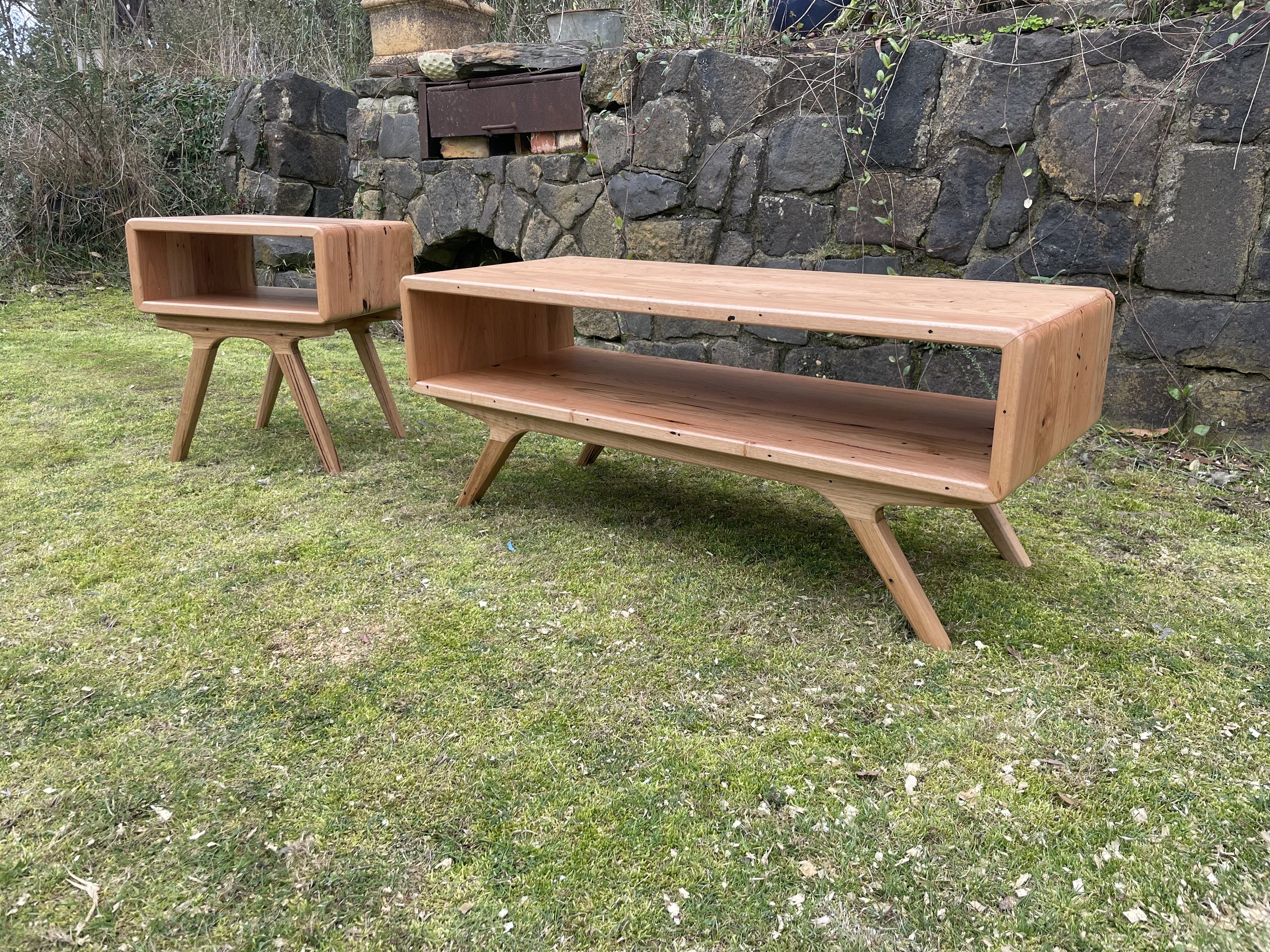 Two wooden tables with slanted legs placed outside on grass in front of a stone wall.