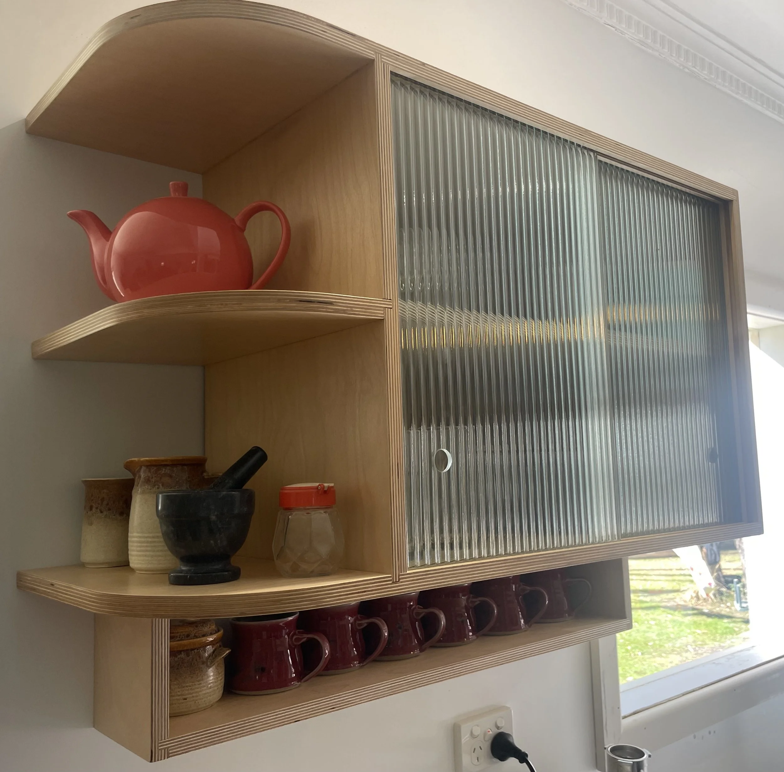 Wooden kitchen cabinet with a metal sliding door, featuring a red teapot on the top shelf, ceramic mugs on the bottom shelf, and various jars and a mortar and pestle on the middle shelf.