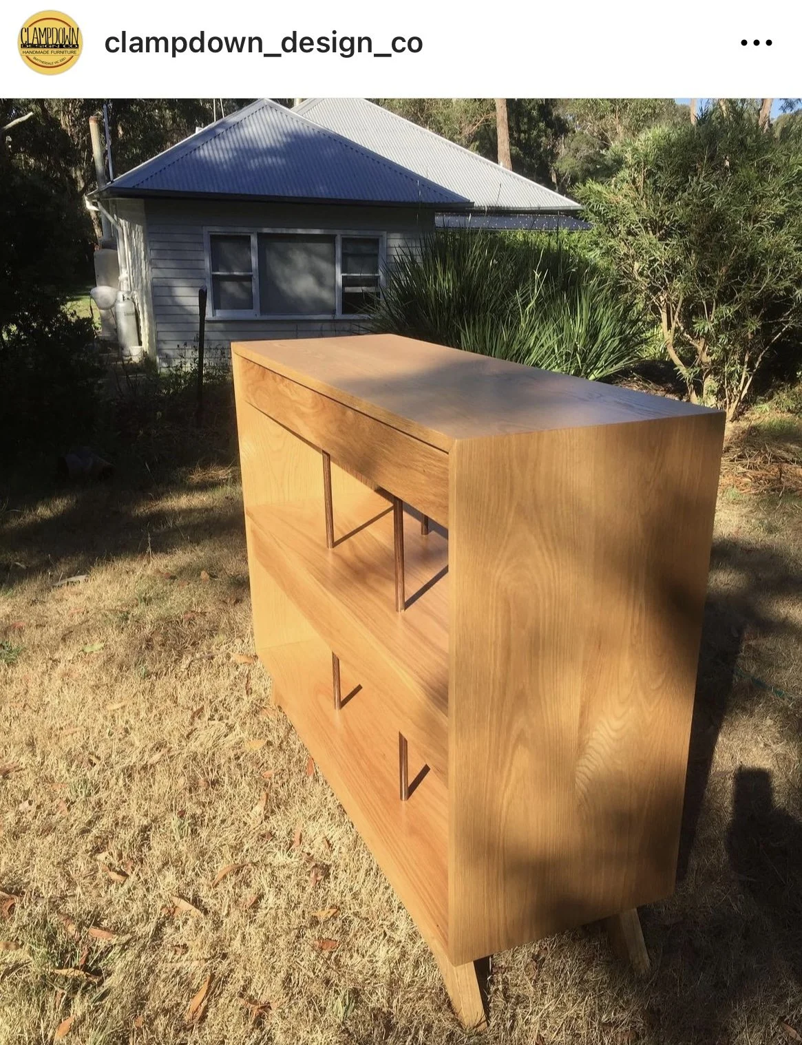 A wooden bookshelf with three shelves, placed outdoors on the grass with a house and trees in the background.