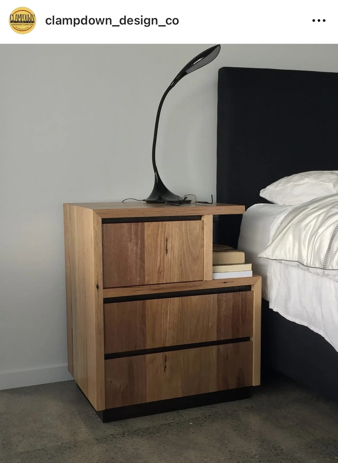Wooden nightstand with three drawers beside a bed with white linens and pillows, topped with a modern black curved desk lamp, against a plain white wall.