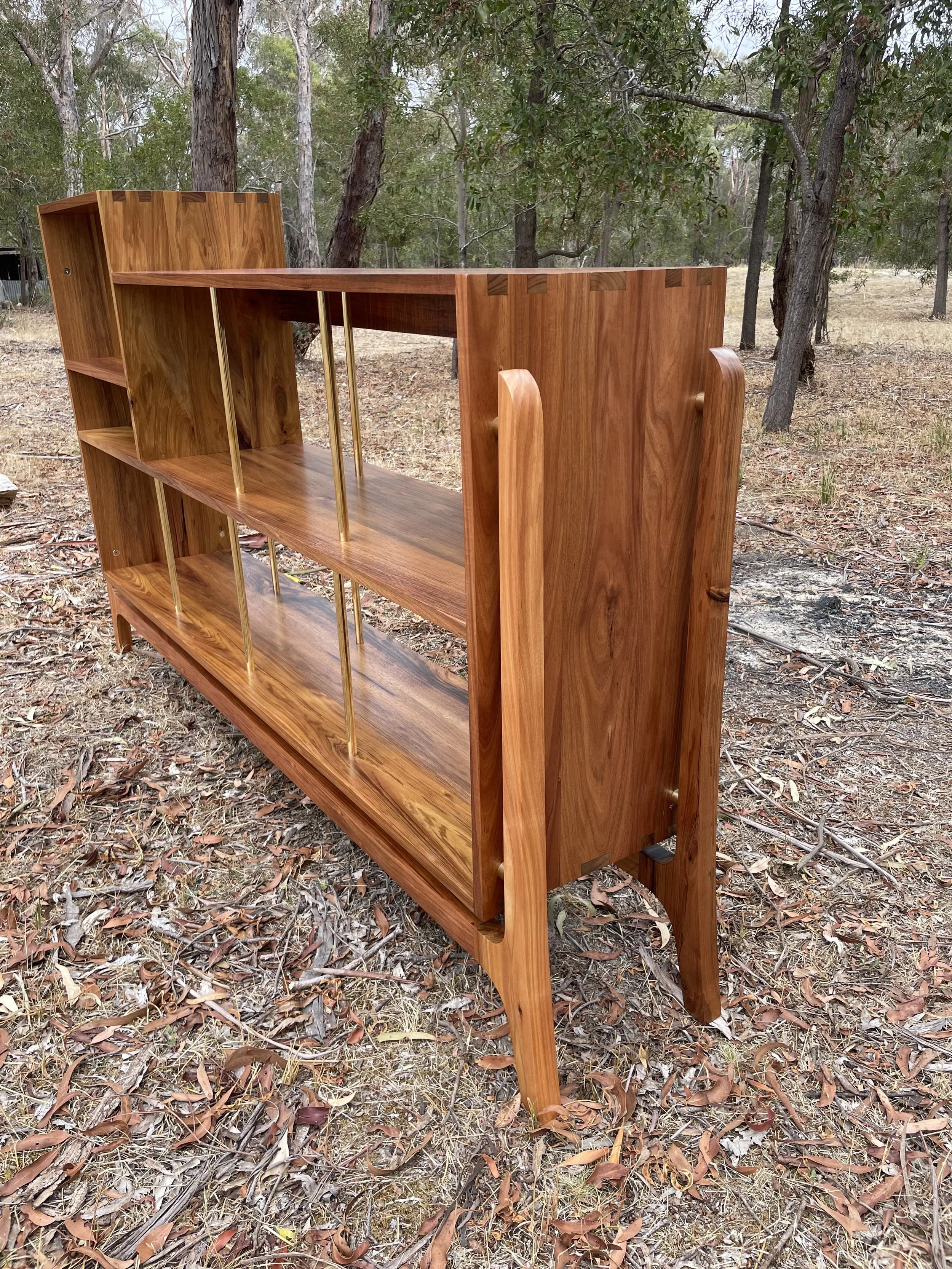 A wooden bookshelf with open sides and shelves, supported by vertical gold-colored metal rods, set outdoors among trees and dry leaves.