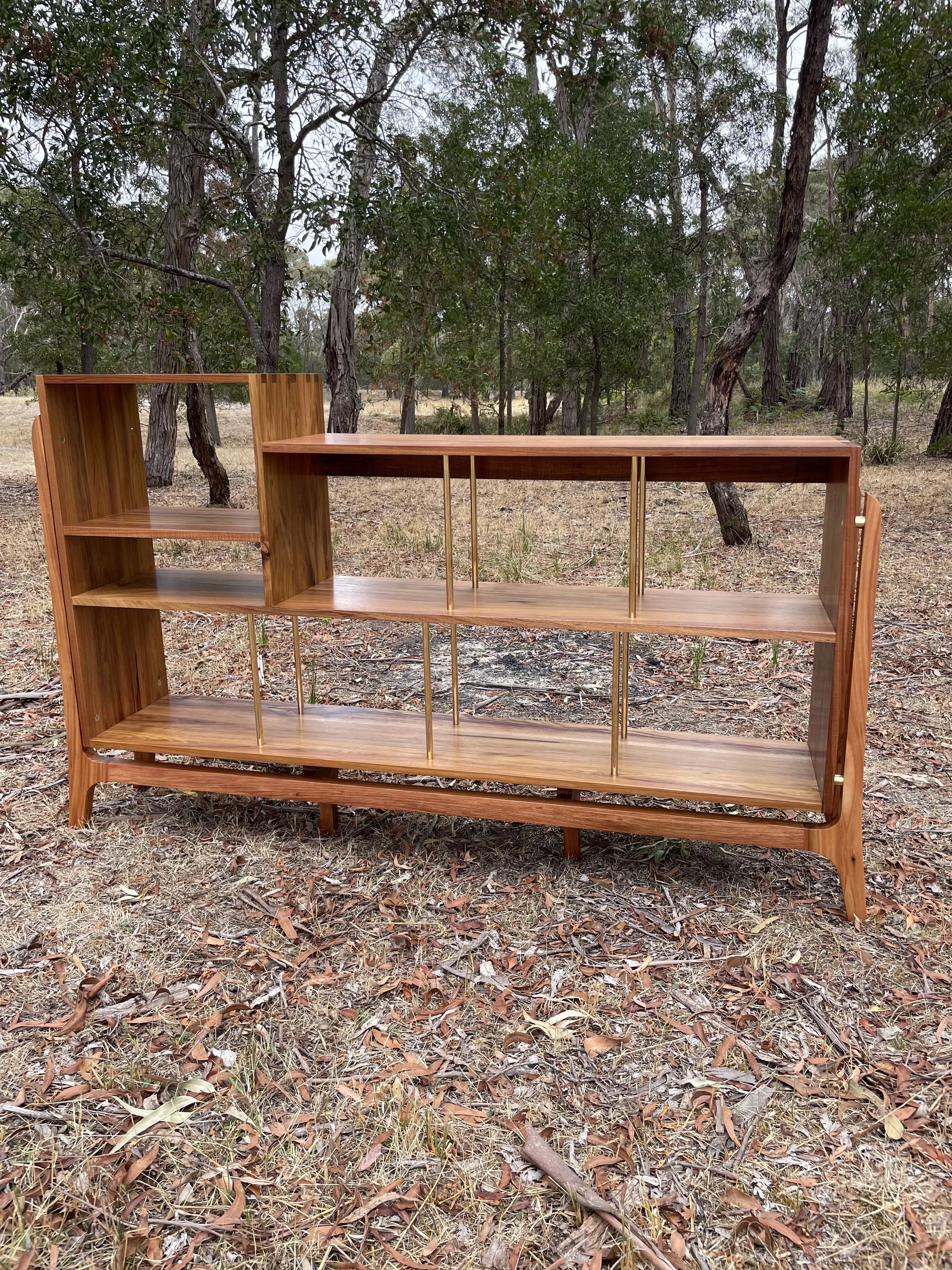A wooden bookshelf with multiple compartments and gold-colored metal supports stands outdoors on a ground covered with dry leaves, with trees and a forested area in the background.