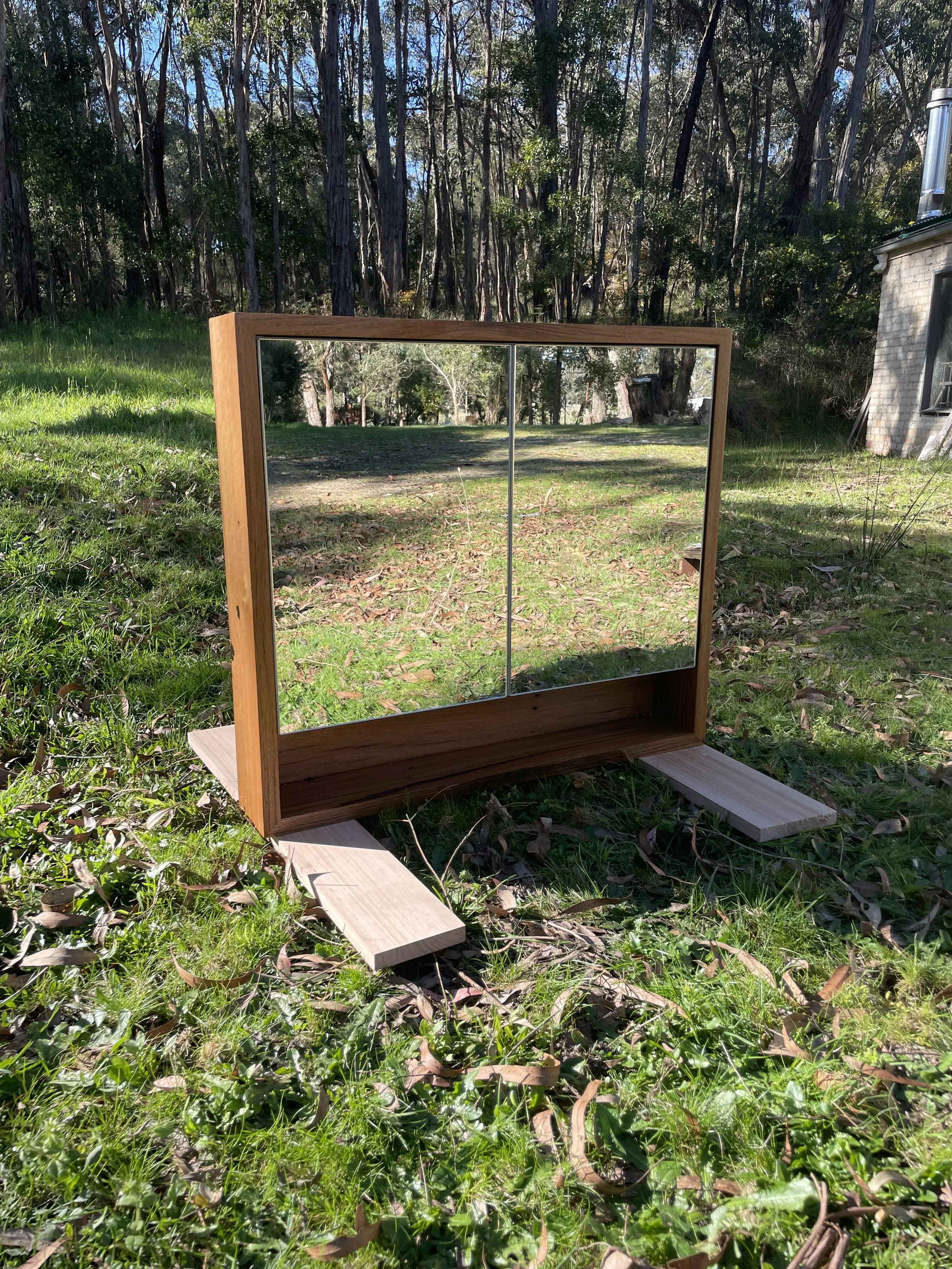 A wooden framed mirror with a double-pane glass is positioned outdoors on a grassy area with trees in the background and a small house to the right.