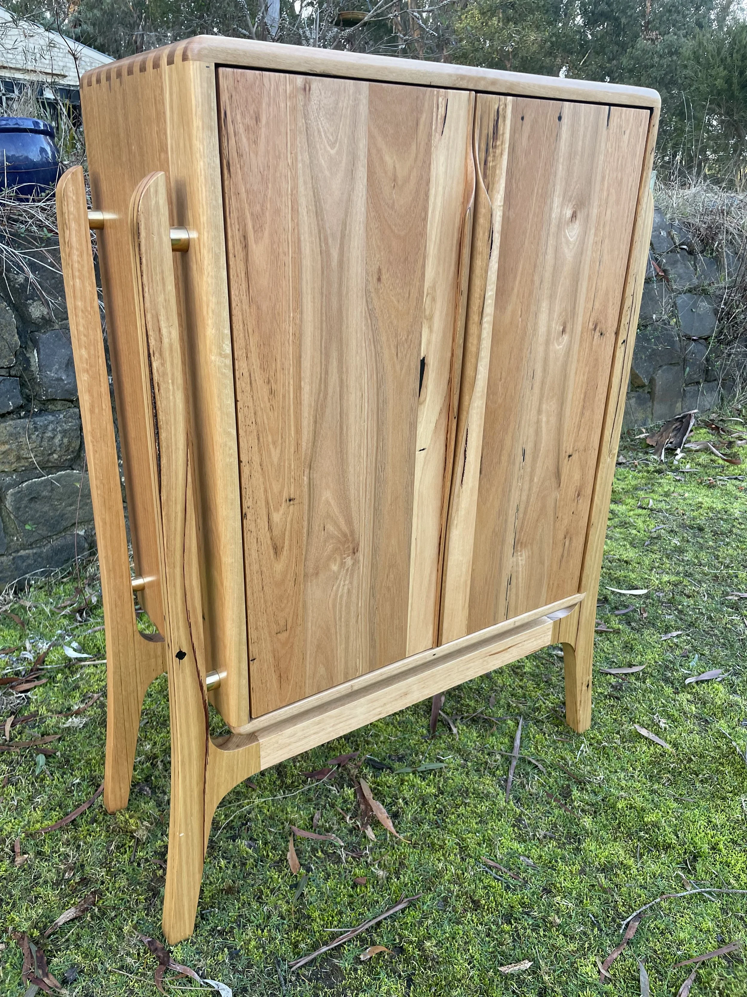 A wooden cabinet with a natural finish, standing outdoors on grass, with a stone wall and trees in the background.