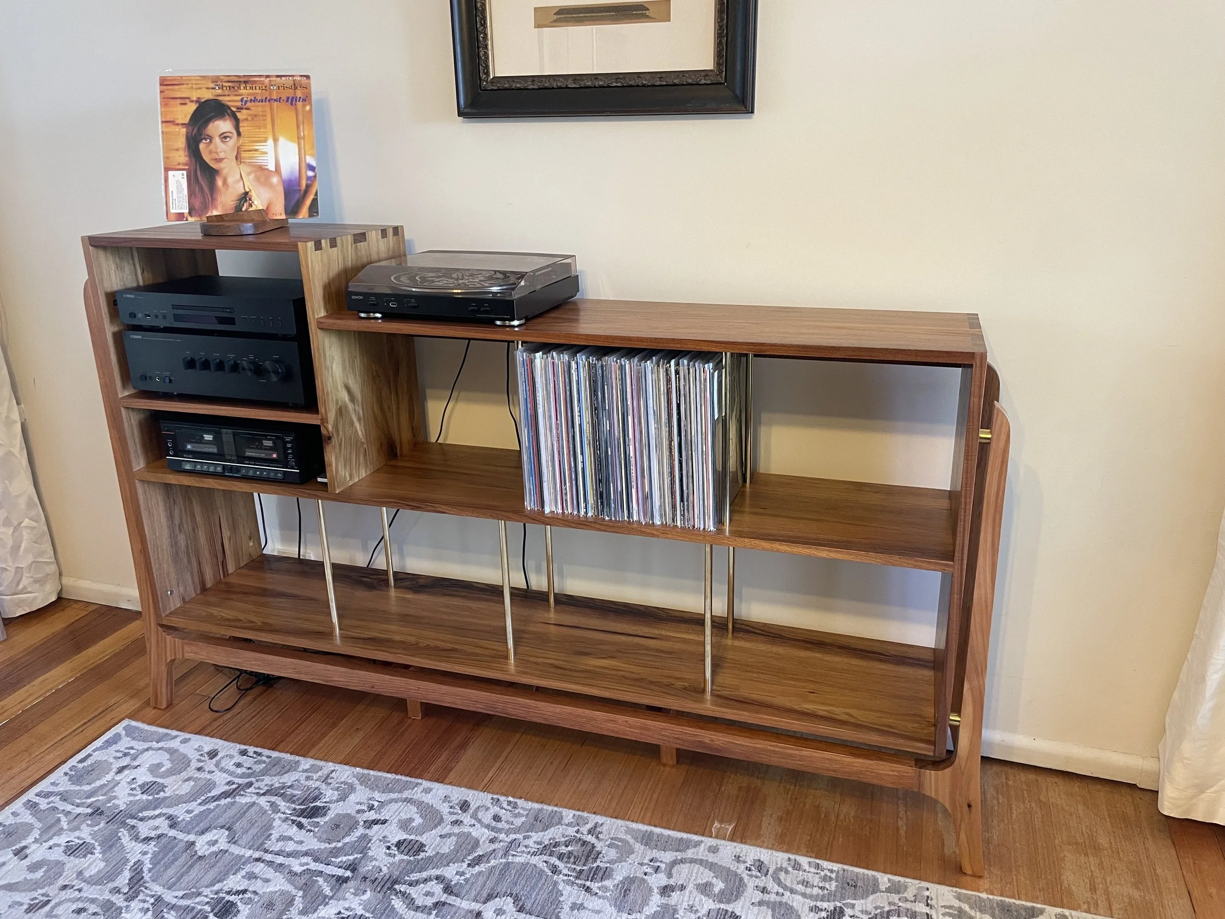 Wooden bookshelf with vinyl records and electronic equipment, a framed picture, and a poster of a woman on top.