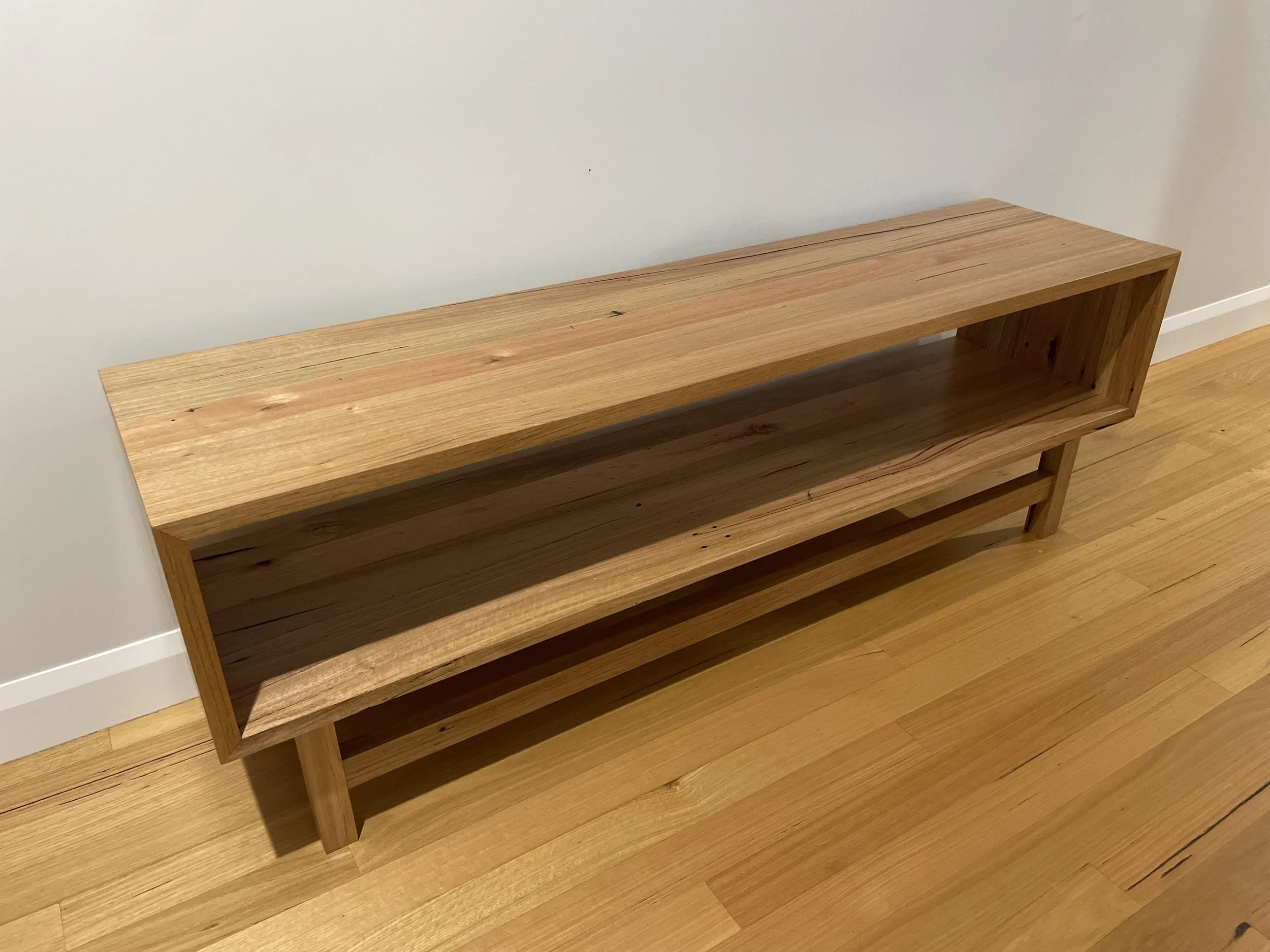 A wooden bench with a rectangular open shelf and a lower rack, placed against a white wall on a hardwood floor.