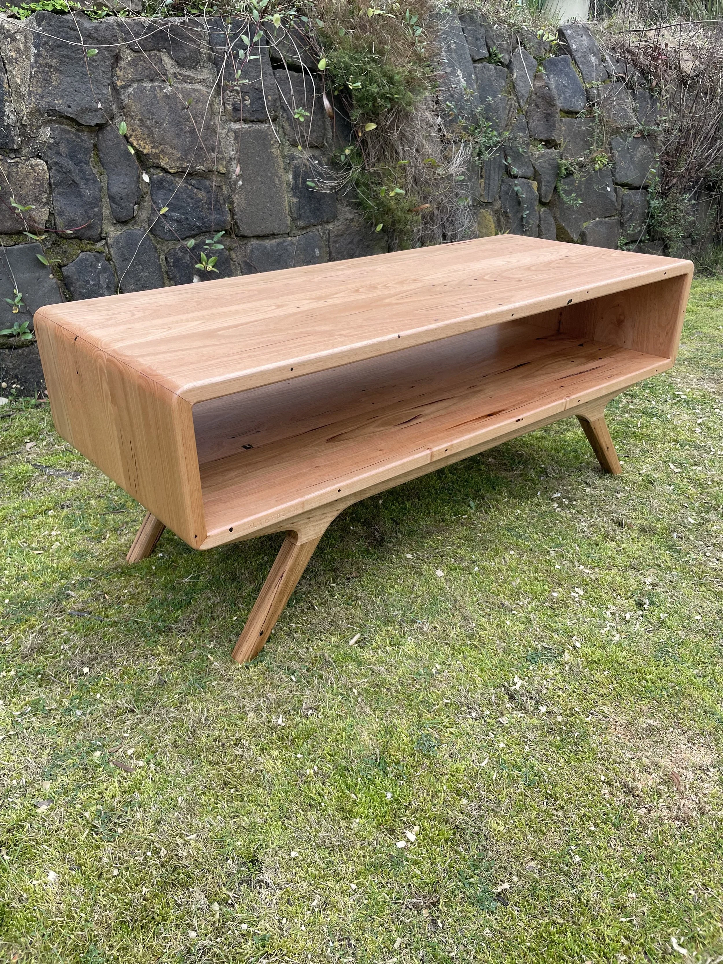 A wooden furniture piece with a rectangular open shelving design, supported by four angled legs, placed outdoors on grass with a stone wall and plants in the background.