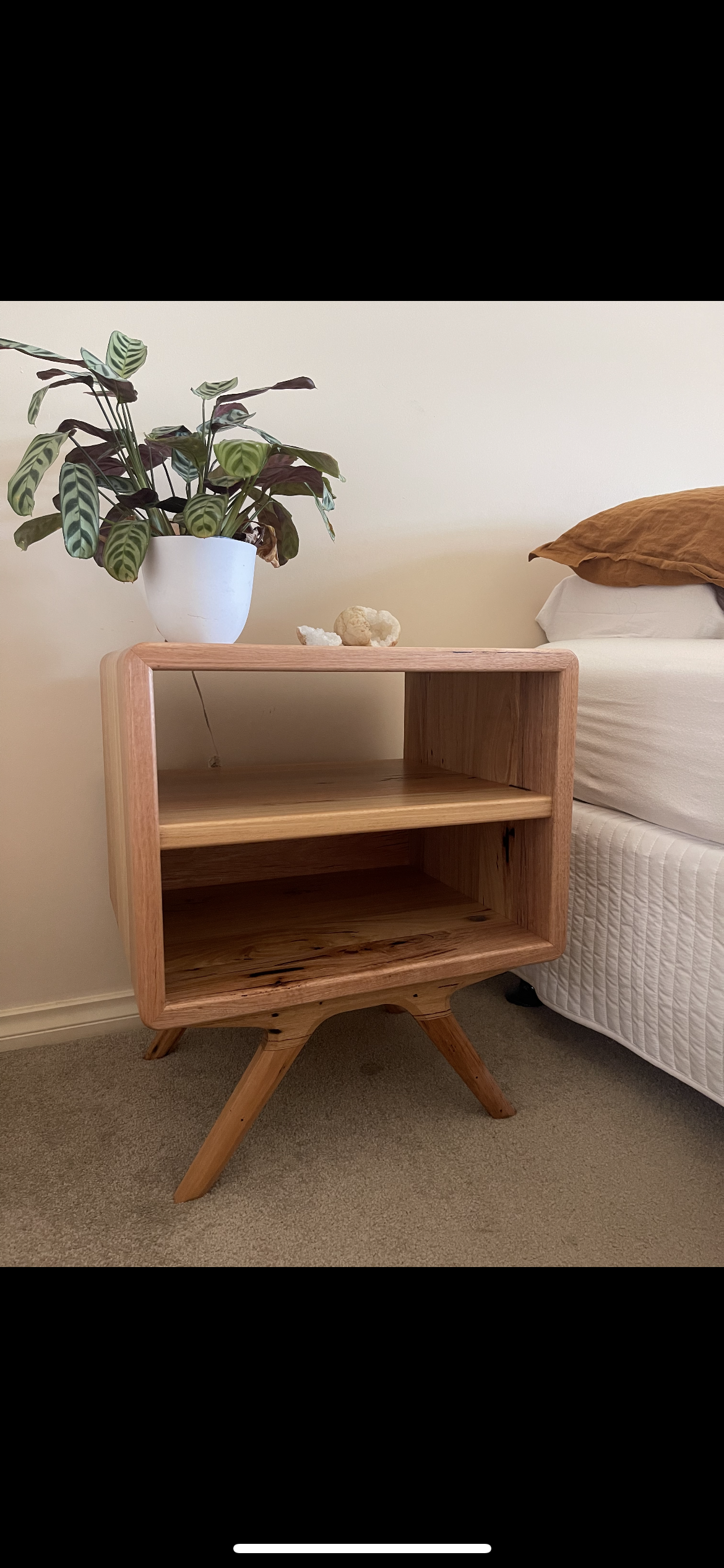 A wooden bedside table with two open shelves, placed next to a bed with white and brown pillows. A potted plant with large green and purple leaves on top of the table, and a small decorative object on the surface.