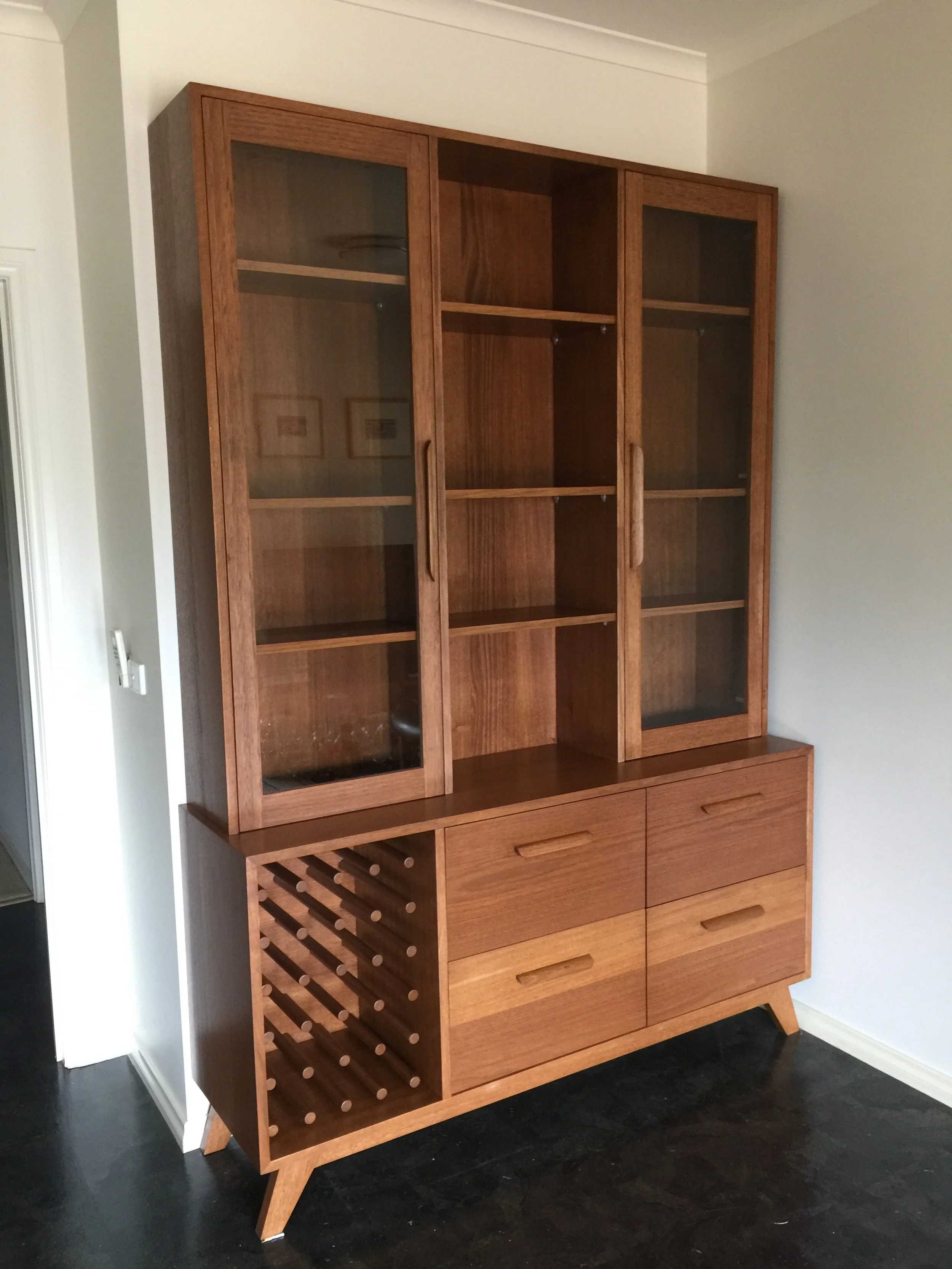 A tall wooden china cabinet with glass doors on the upper section and four drawers and a wine rack on the lower section, standing on angled legs in a room with dark flooring and light-colored walls.