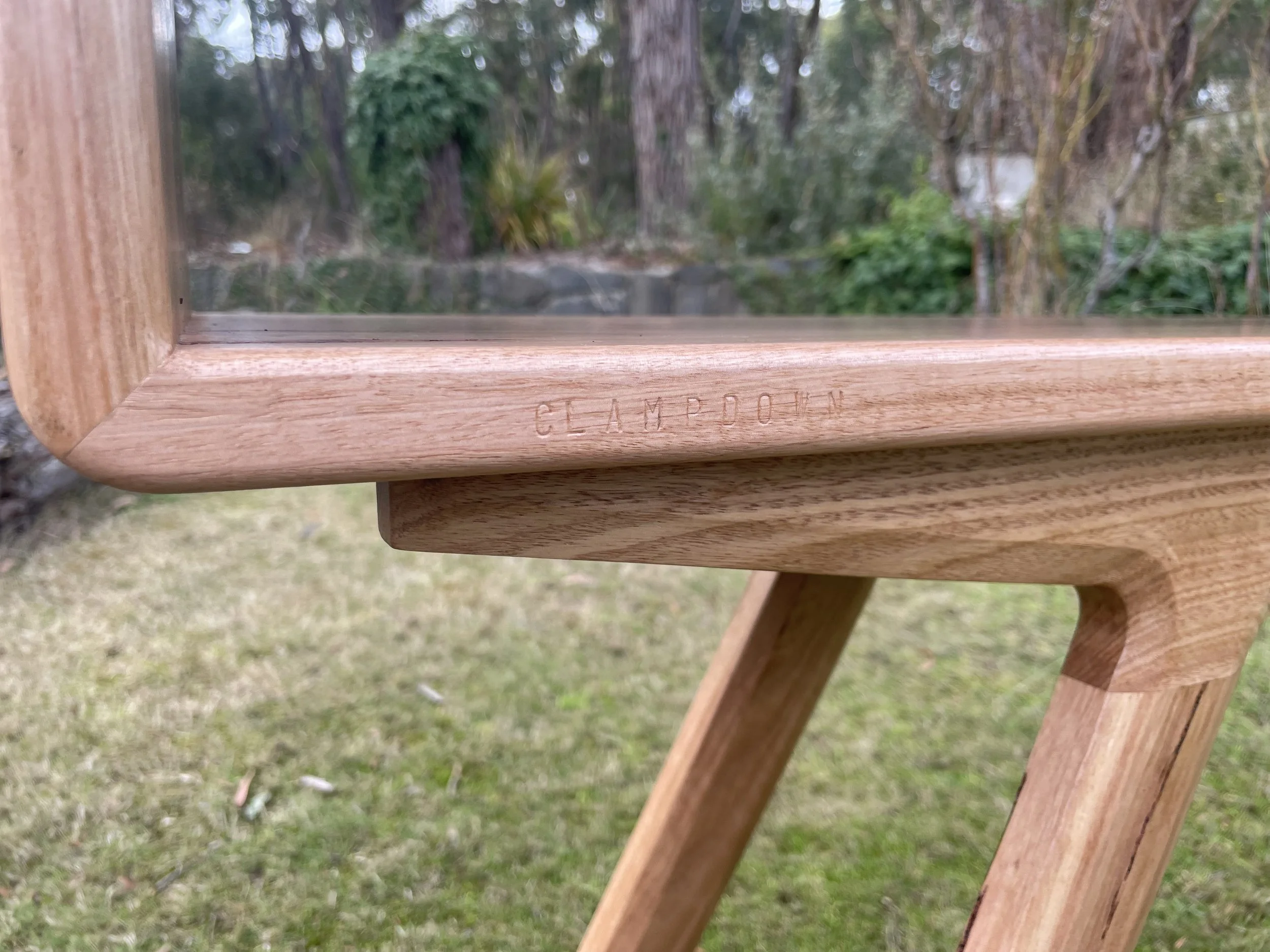 Close-up of a wooden table leg and the edge of the tabletop, with the brand name 'GLEMPTON' faintly engraved on the side of the wood.