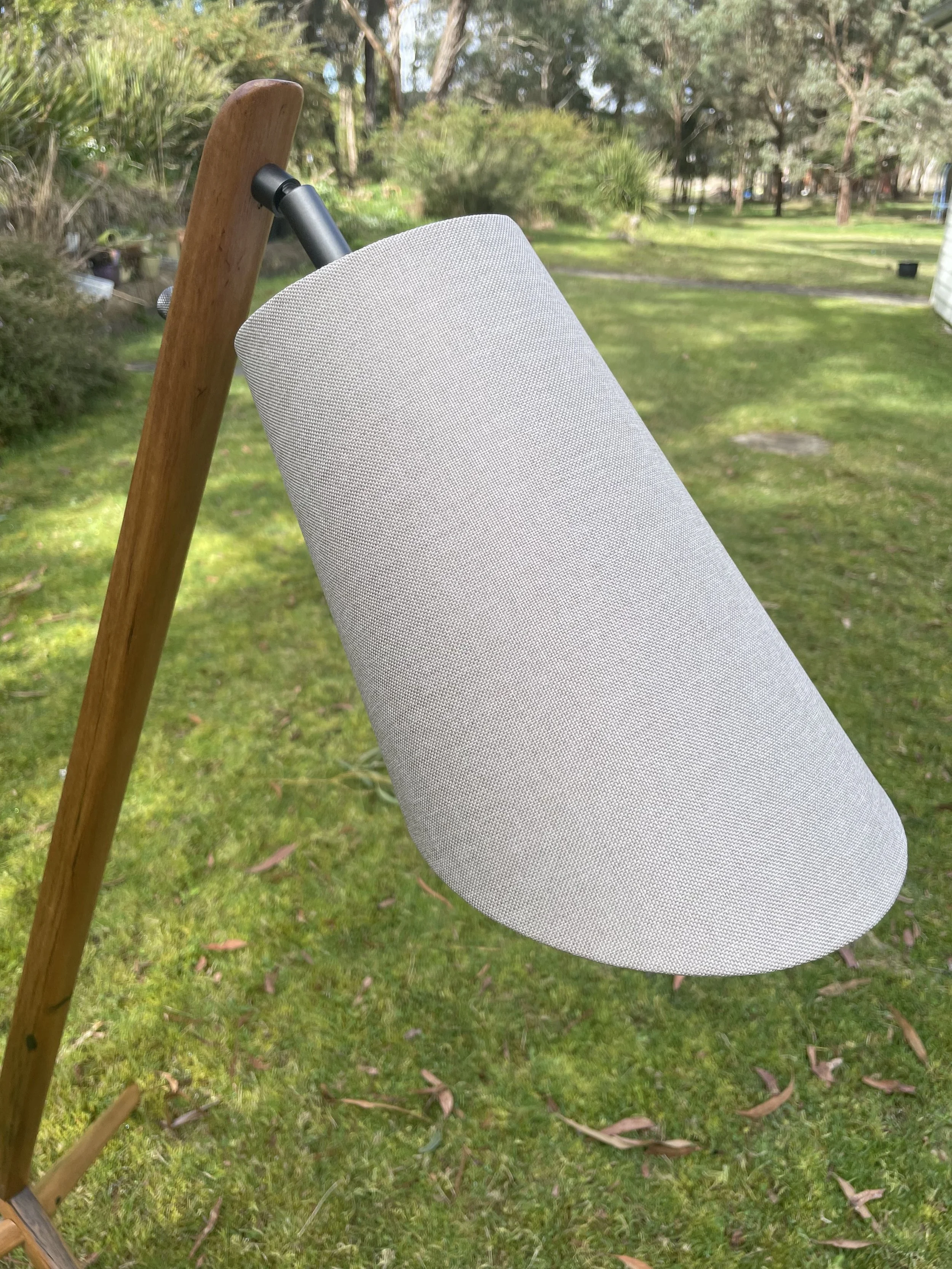 Close-up of a modern speaker with a gray fabric exterior, attached to a wooden stand, outdoors on a grassy yard with trees in the background.