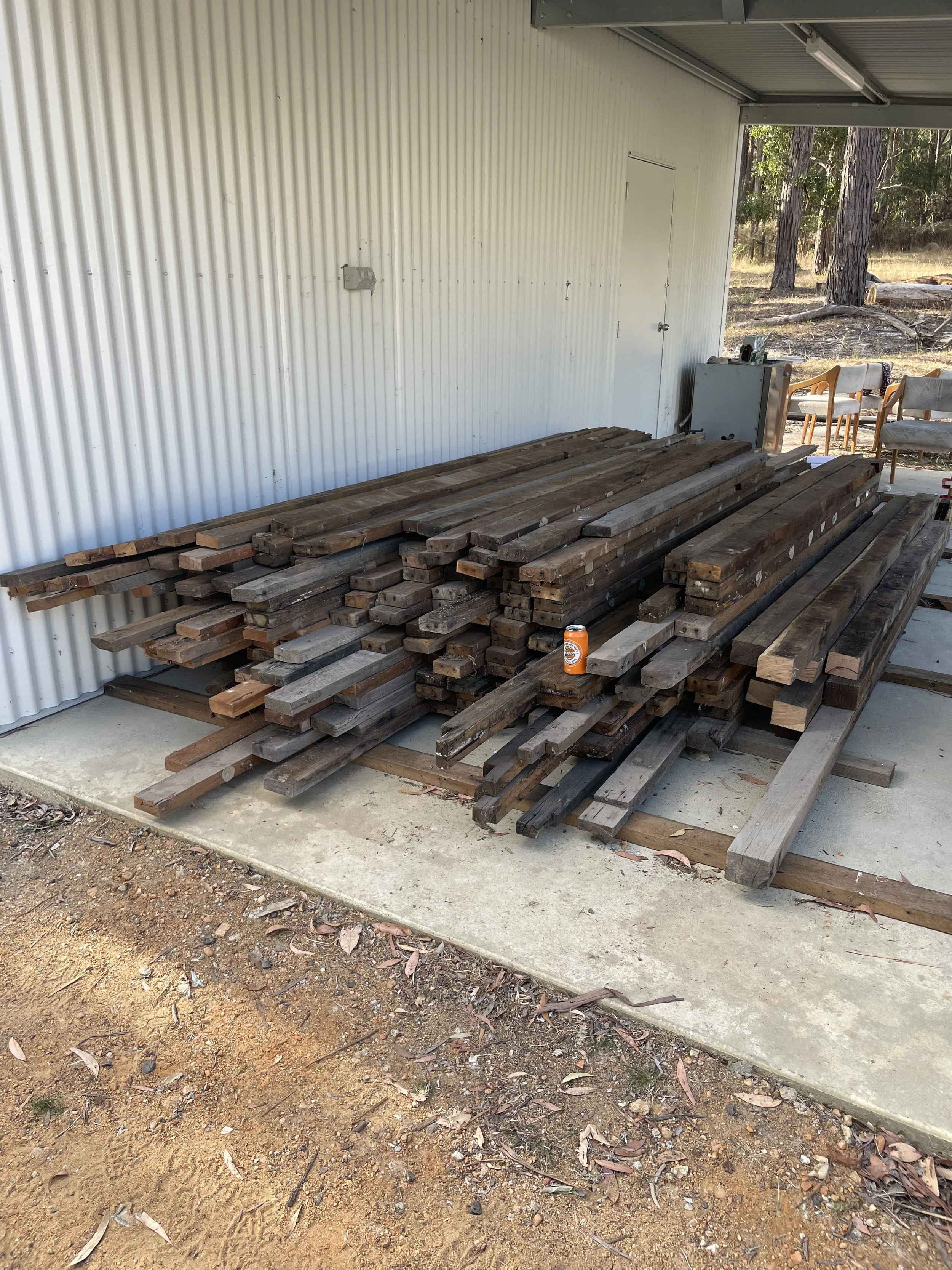 A large pile of weathered wooden planks stacked outside a building with a corrugated metal wall. There is a concrete slab underneath and a dirt area with leaves in front. In the background, there are trees and outdoor furniture.
