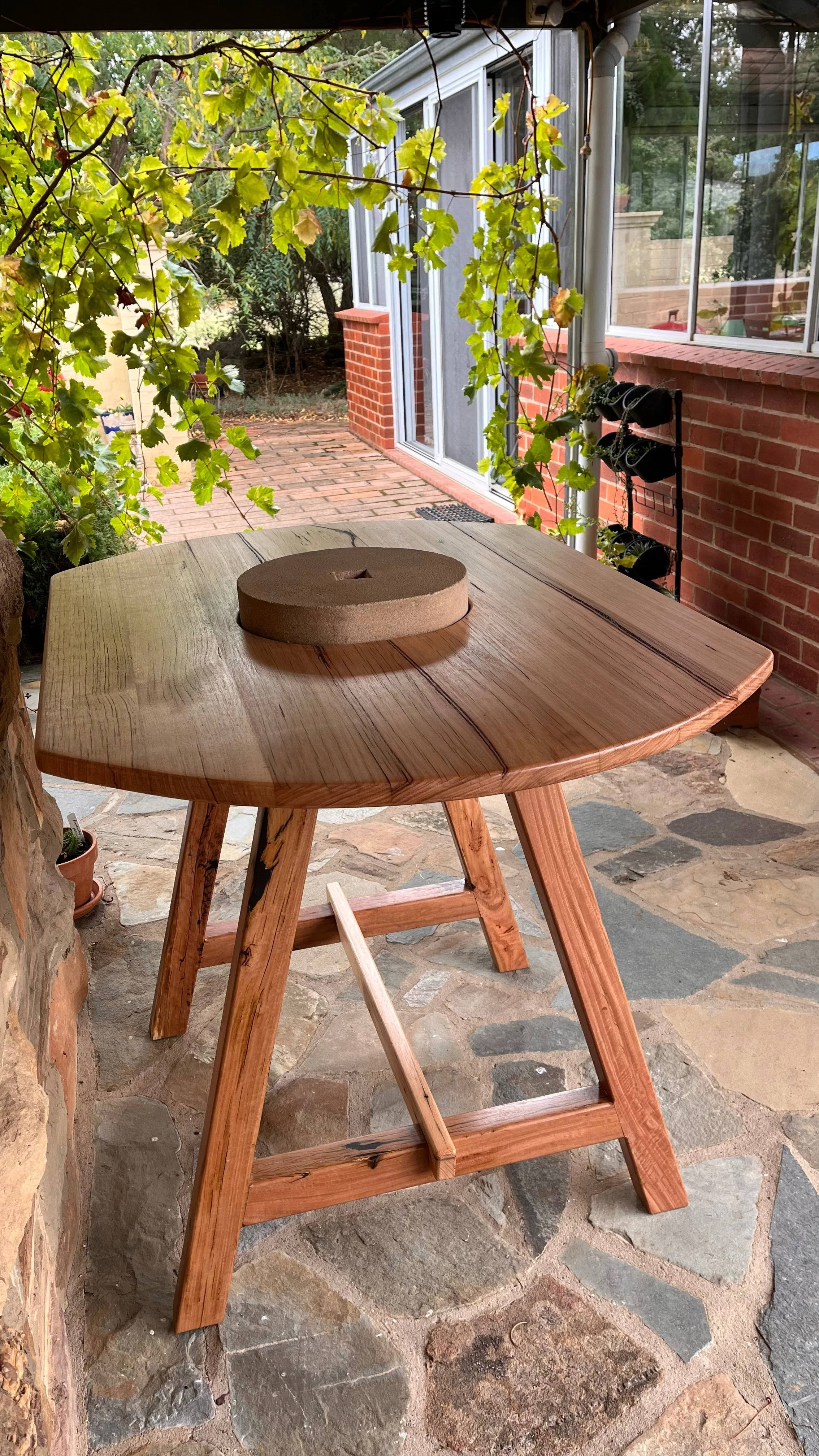 A wooden table with a saucer on top, set on a stone patio with foliage and a brick and glass house in the background.