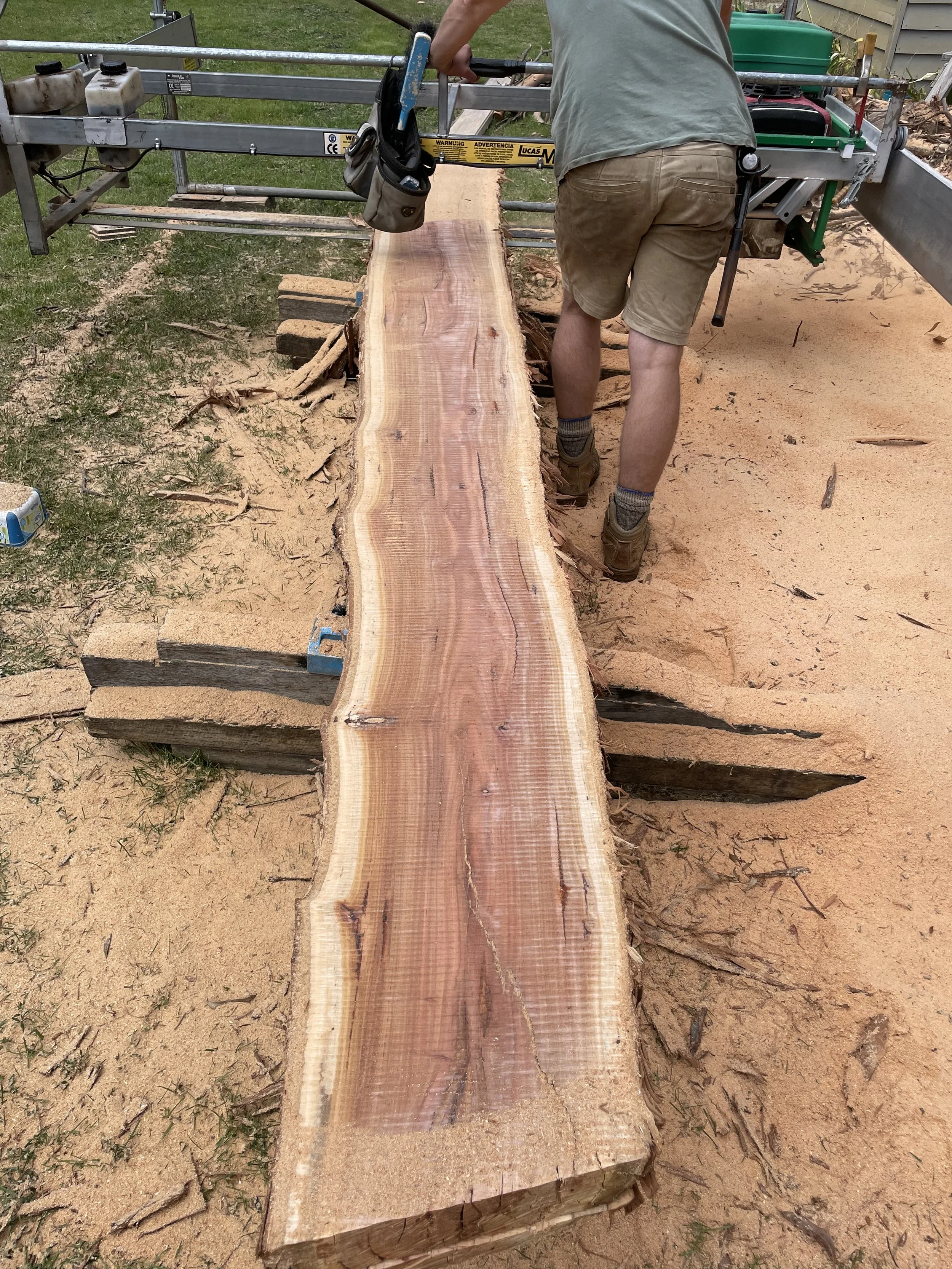 A person working on a large, flat wooden slab on a sawmill, with sawdust and wood pieces around, outdoors on a dirt surface.
