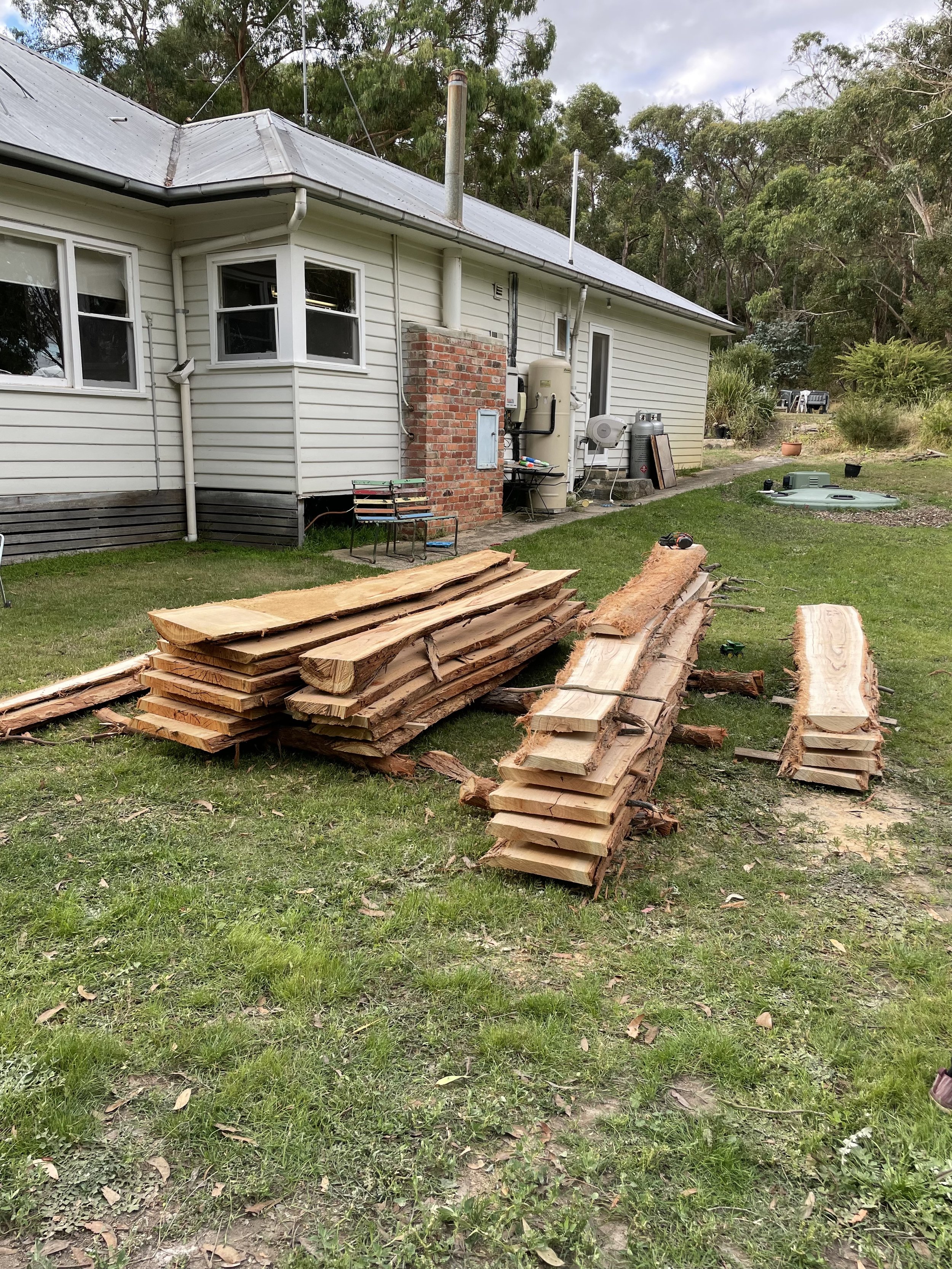 Stacks of cut and split wooden planks laid on the grass in the backyard of a house, with a house visible in the background.
