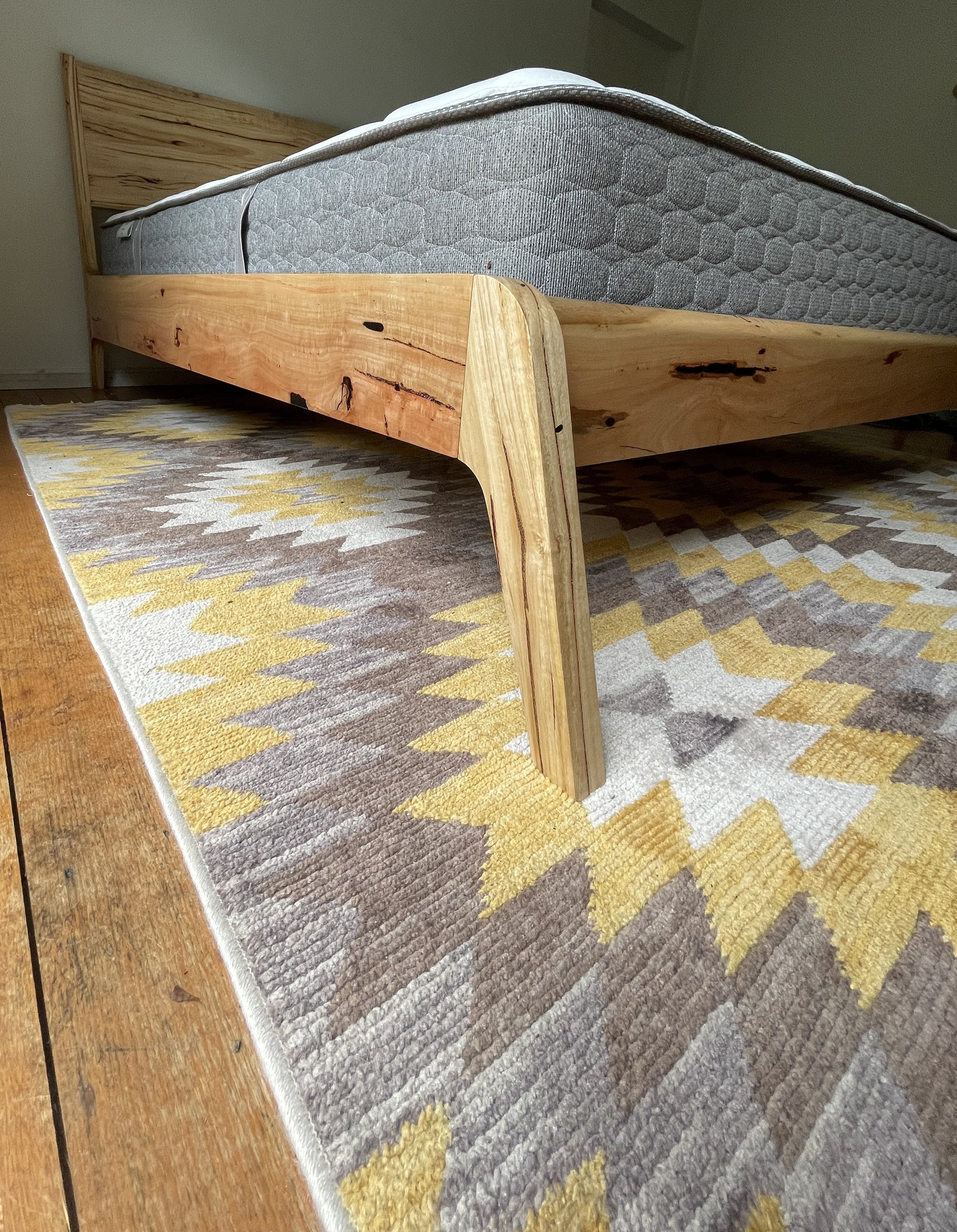 Close-up of a wooden bed frame with a natural finish, placed on a geometric patterned area rug.
