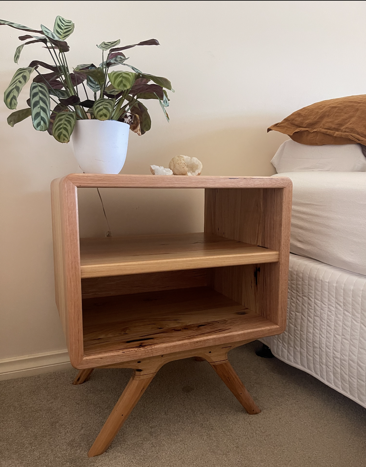A wooden nightstand with two open shelves, standing next to a bed. The nightstand has angled legs and a natural wood finish. On top, there is a potted plant with green and purple patterned leaves, and a sculpture or decorative object.